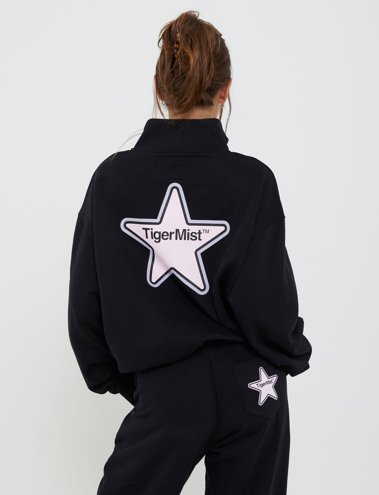 A person with long hair tied up is seen from the back, wearing a black jacket and the DELFINA PANT - BLACK from TIGERMIST, adorned with white star patches. The jacket features a large star with "TigerMist™" written inside, highlighting the brand TIGERMIST. The pants sport a smaller star patch on the right pocket.