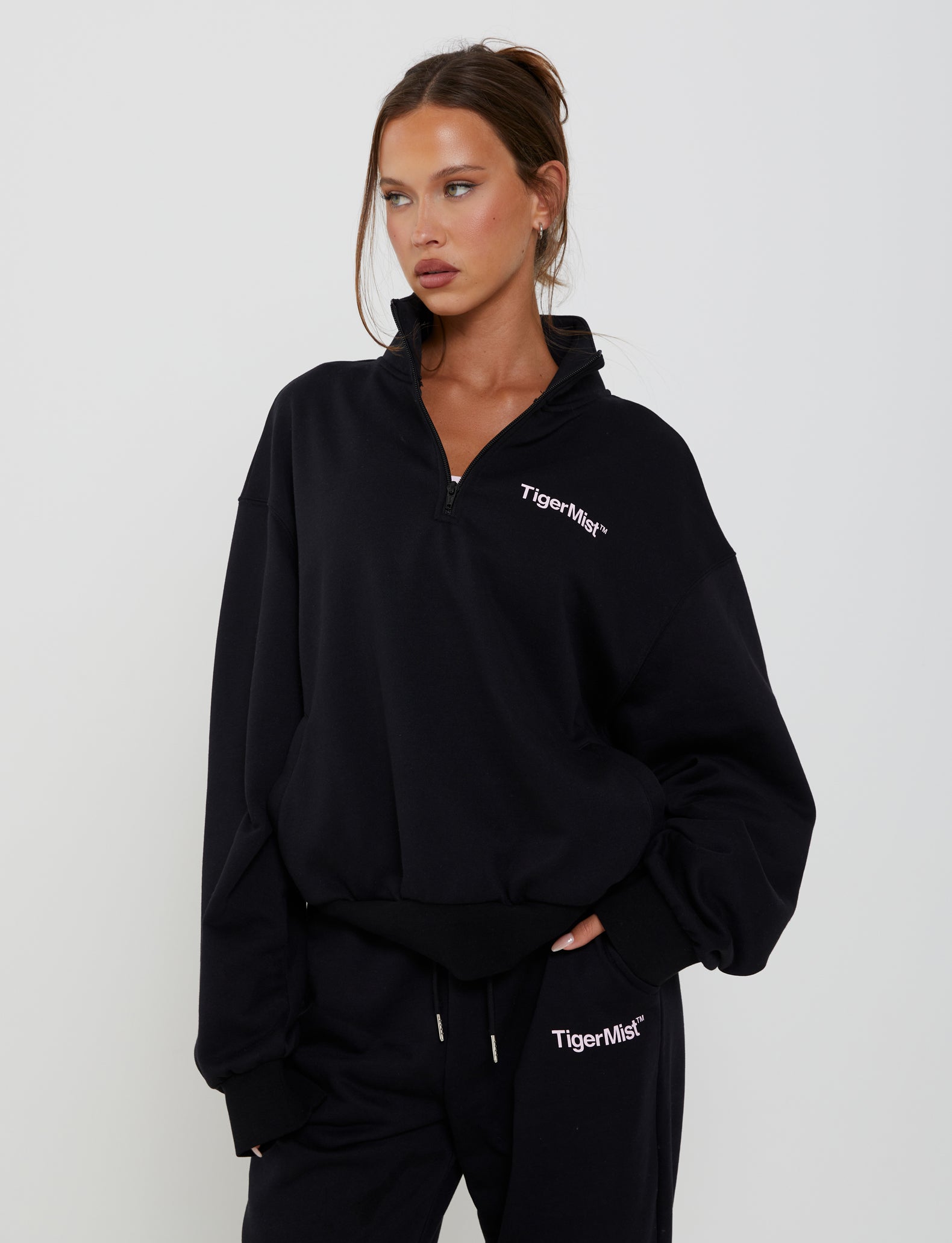 A woman stands against a plain background wearing the TIGERMIST brand DELFINA PANT in black, with her hair tied back and one hand in her pocket. She gazes off to the side.