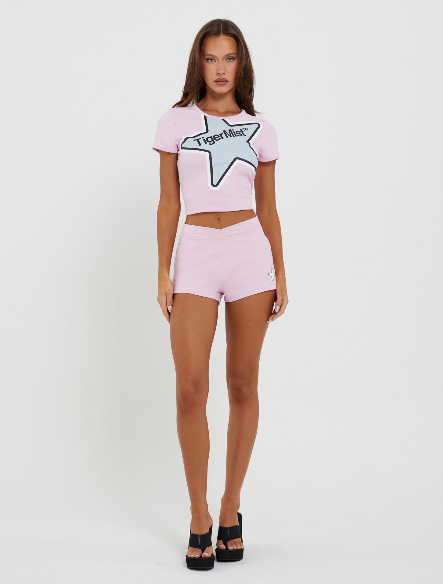 A person with long hair stands against a white background, dressed in a light pink two-piece outfit featuring the TIGERMIST DELANEY TEE - PINK with a star design and the text "Tiger Mist," paired with matching fitted silhouette shorts. They complete the look with black high-heeled sandals.