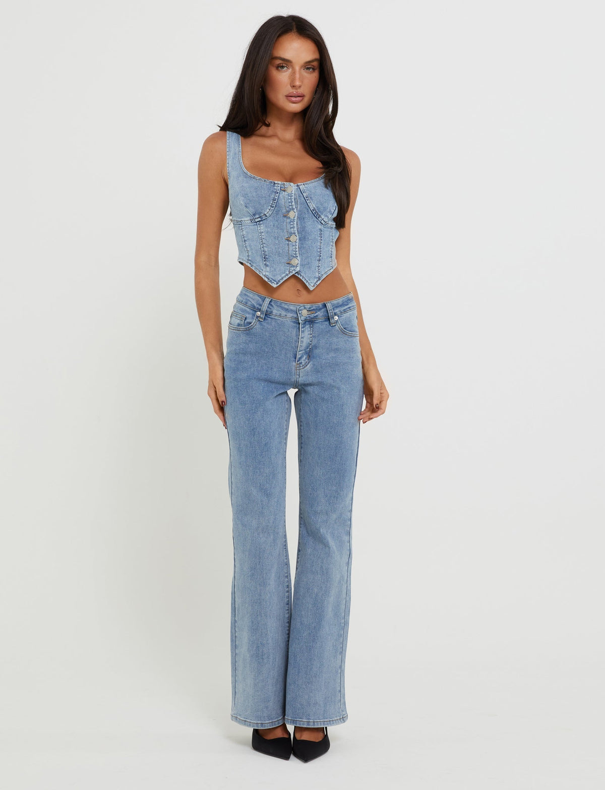 A woman with long, dark hair is posing against a plain white background, wearing light blue denim clothing from TIGERMIST. She has on the TATYANA TOP in light wash, a sleeveless cropped denim top, paired with high-waisted flare jeans and black pointed-toe heels.