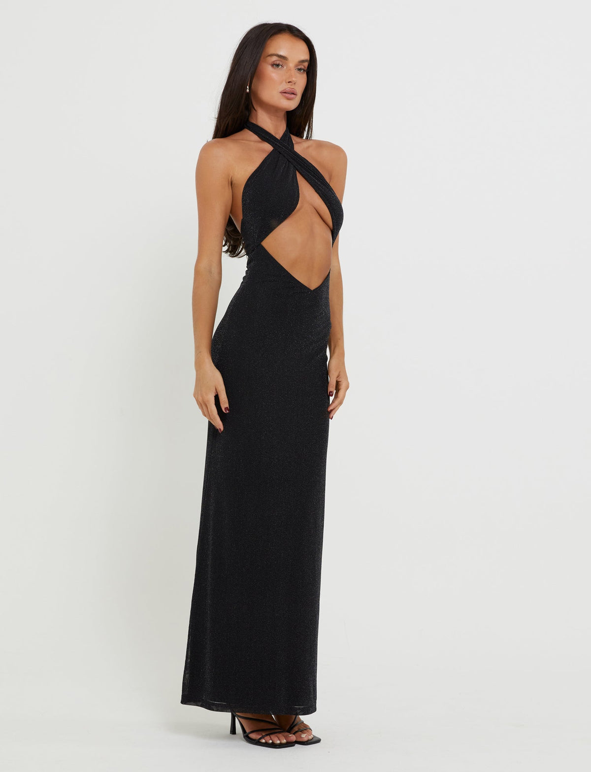 A woman wearing the TIANAH MAXI DRESS - BLACK from TIGERMIST stands against a plain white background. She has long dark hair and is sporting black high-heeled sandals. Posing with one hand resting on her hip, she looks directly at the camera.