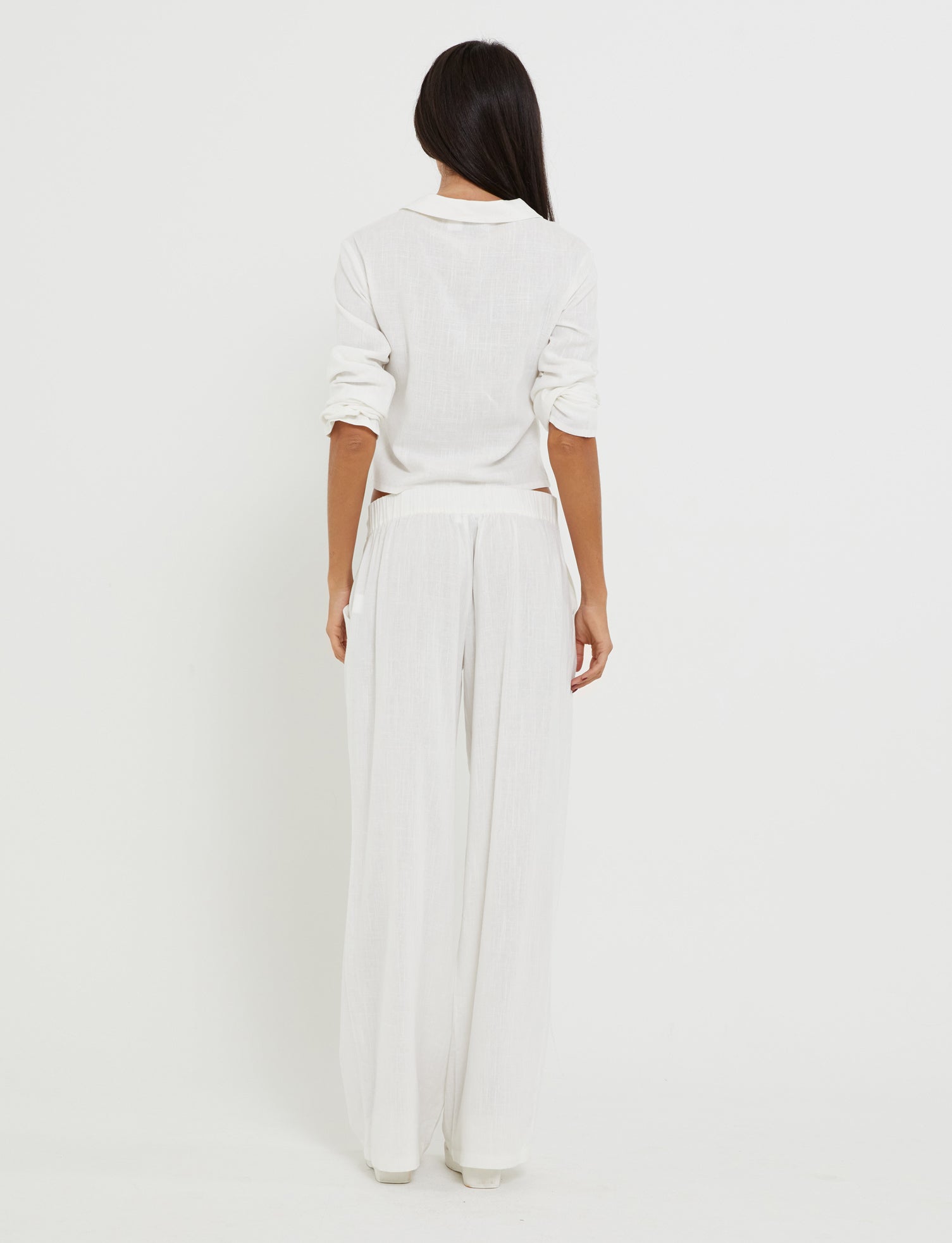 A person with long dark hair stands facing away, wearing the AIDEN TOP - WHITE from TIGERMIST paired with loose white pants. The outfit is relaxed and minimalist, featuring rolled-up sleeves and a slightly untucked hem. The background is completely white, creating a clean and simple look.