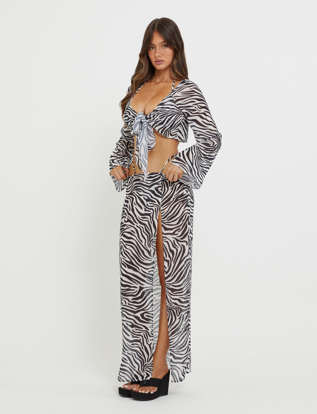 A woman stands against a plain white background wearing a striking Tiger Mist ensemble. The outfit features the AYESHA SKIRT - BLACK : ZEBRA, a high-slit maxi skirt, paired with a long-sleeved, tie-front crop top adorned in zebra print. She completes the look with black platform sandals. Her long hair cascades loosely around her shoulders, and she has a neutral expression on her face.