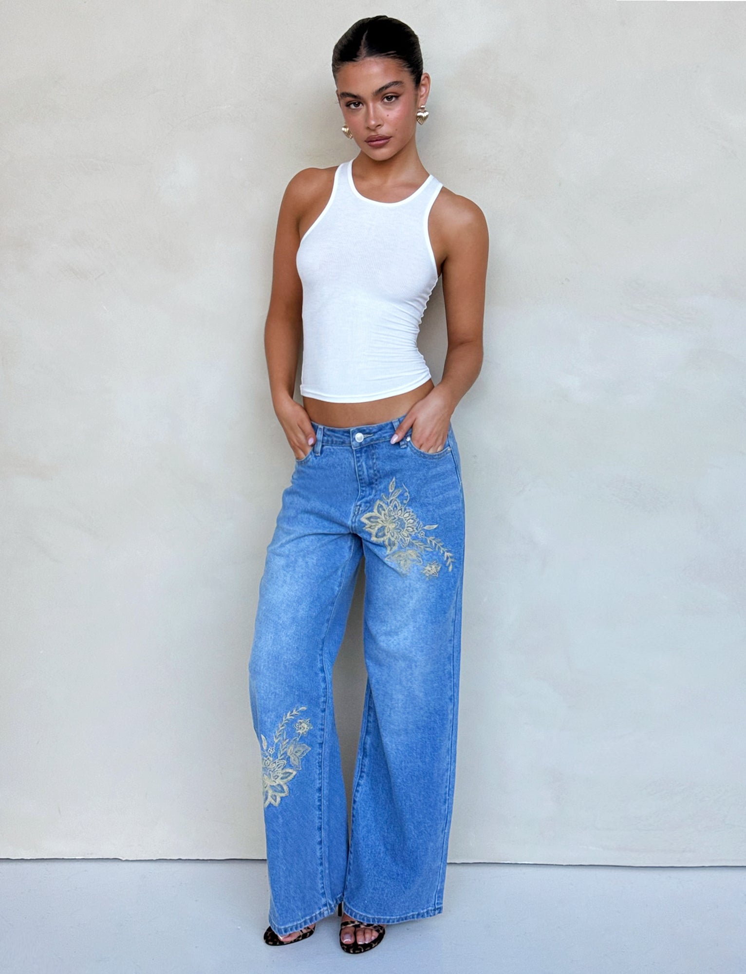 A person with dark hair pulled back is wearing a fitted white tank top and the VEGA JEAN - BLUE by TIGERMIST, featuring mid-blue denim with signature gold floral embroidery on the thighs. They are standing against a plain beige background with their hands in their pockets and are wearing black heeled sandals.