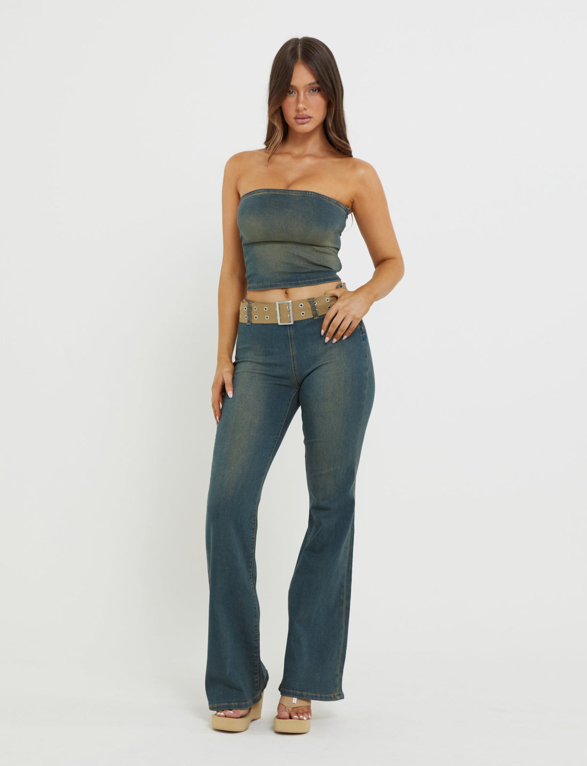 A woman stands against a white background wearing the TIGERMIST ISABEL JEAN - BLUE denim jumpsuit, paired with a tan belt featuring metal eyelets. She accessorizes with tan platform wedge shoes. Her long, straight hair is parted in the middle and cascades over her shoulders.