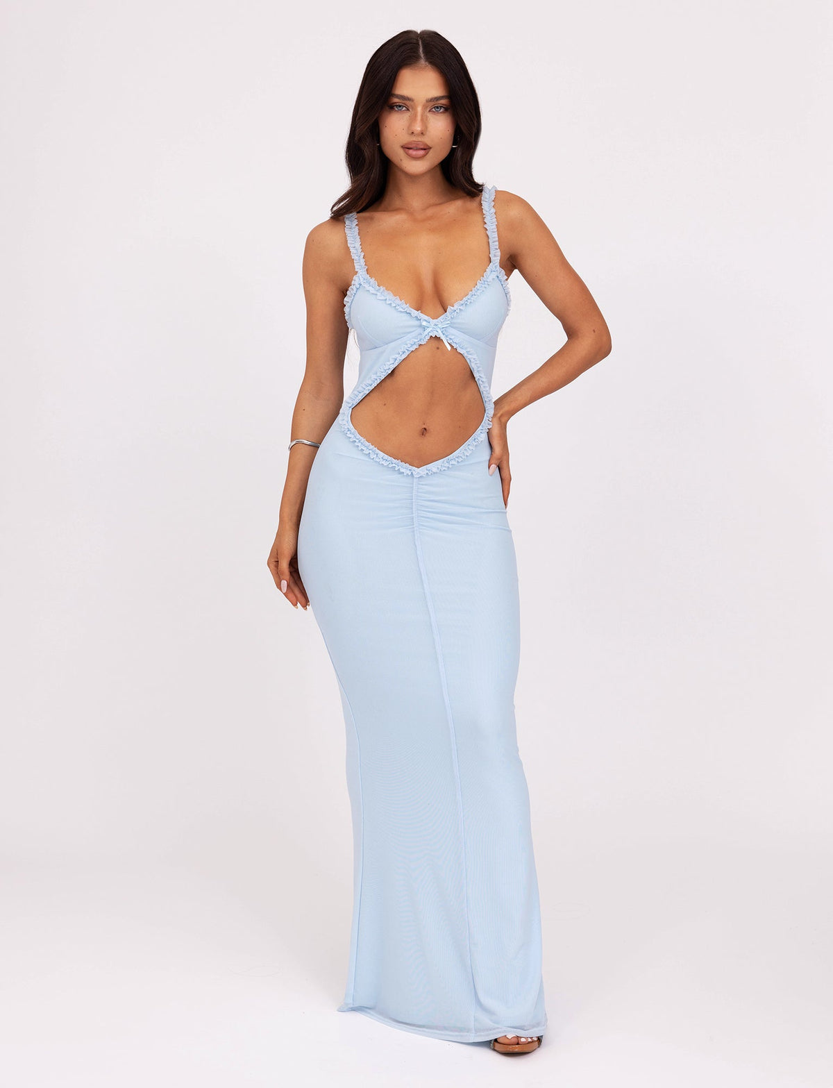 A woman with long, dark hair poses against a white background, wearing the light blue floor-length RAYLA DRESS - BLUE by TIGERMIST, featuring cut-out sections at the torso and midriff. She has her right hand resting on her thigh and her left hand on her hip.