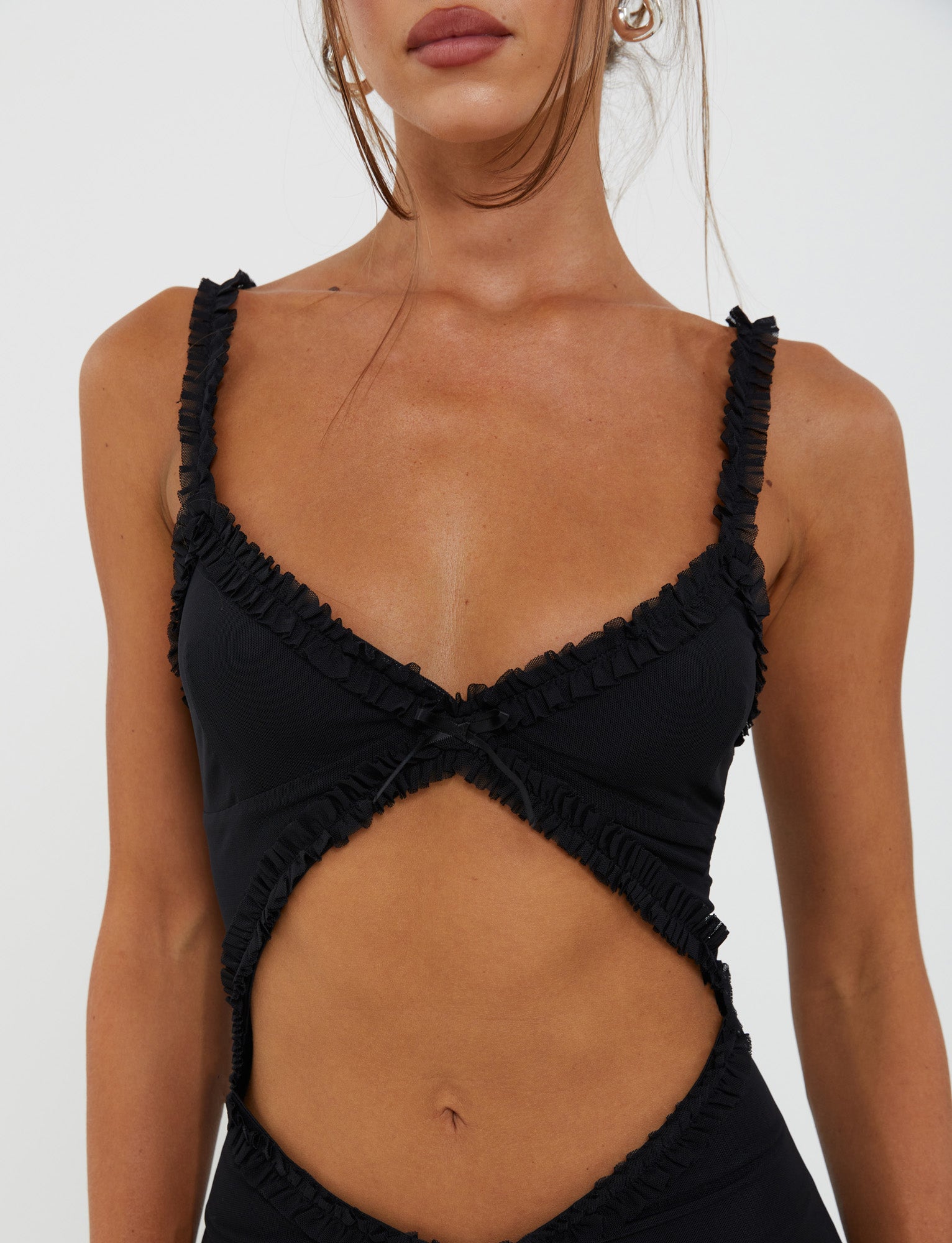 A woman is wearing the Rayla Dress in black from TigerMist, featuring a deep V neckline and cut-out midsection with crisscross detailing on the chest. Her hair is tied back, with tendrils framing her face. Only her upper body and part of her face are visible against a plain background.