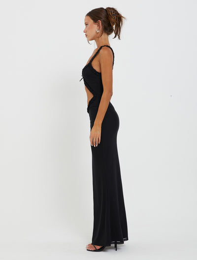 A woman stands in profile against a white background, wearing the RAYLA DRESS - BLACK from TIGERMIST. The form-fitting black dress features cut-out details on the side and a long, flowing skirt. Her hair is styled in an updo, and she accessorizes with earrings and high heels.