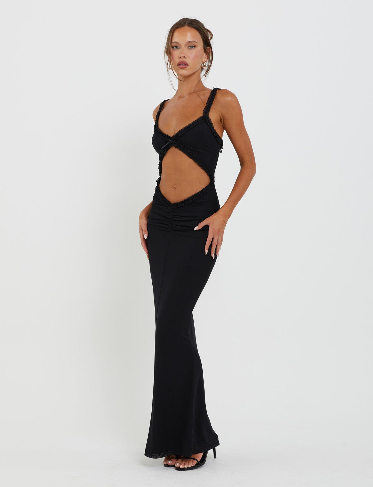 A woman stands poised against a plain background, adorned in the long black RAYLA DRESS by TIGERMIST, featuring cutout details on the sides and back. Her hair is tied up, she wears minimal jewelry, and completes the look with black heels, one hand on her hip and the other by her side.