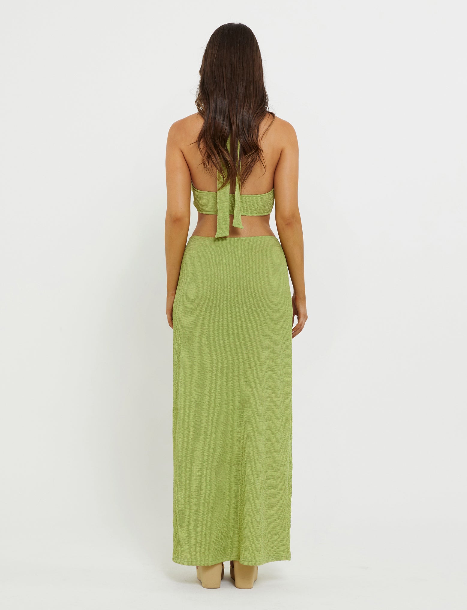 A woman with long dark hair stands facing away from the camera. She is wearing the NEERA TOP - GREEN in lime green from TIGERMIST, paired with a matching long skirt. The halter top features cross-back straps that tie in the middle of her back. She is standing against a plain white background.