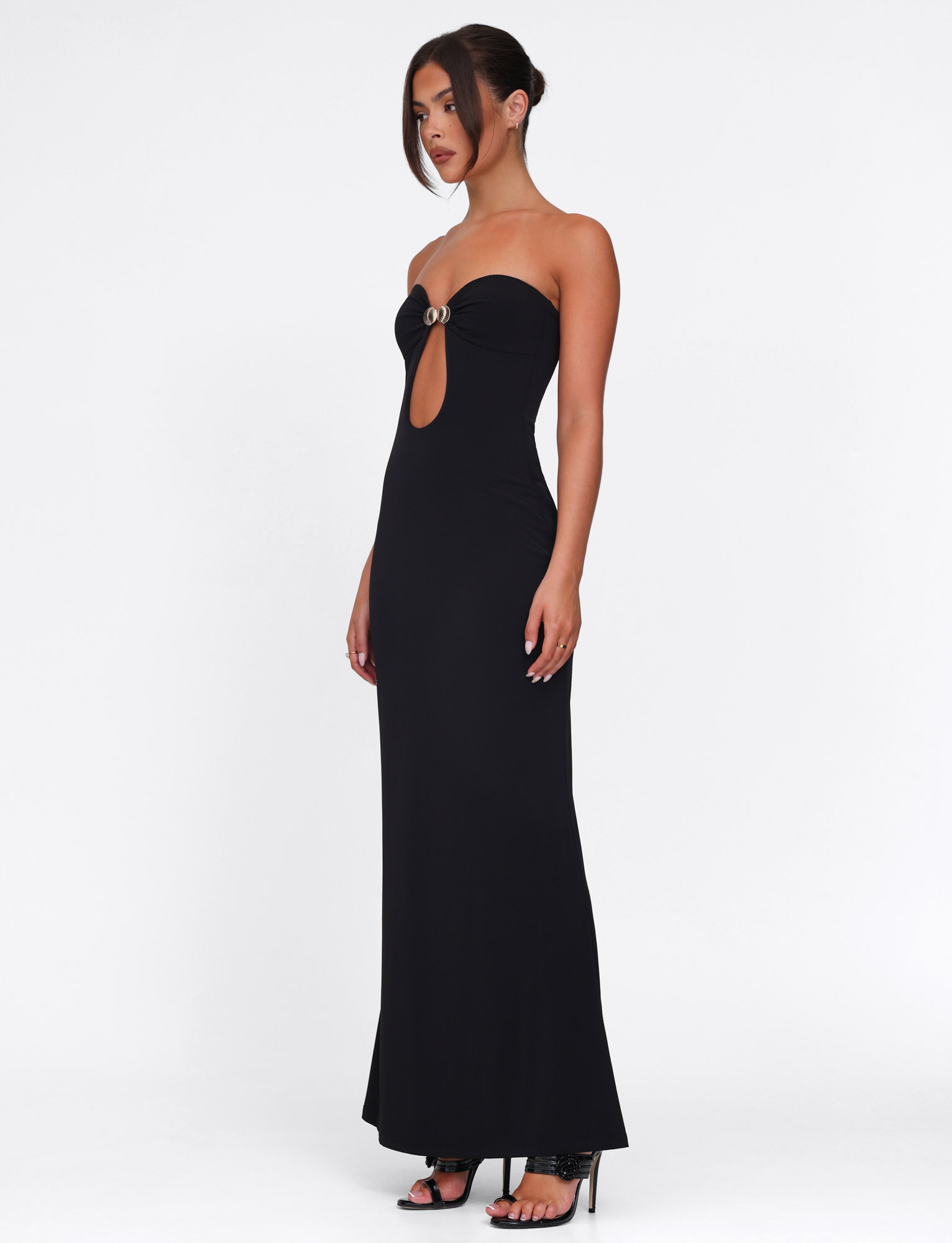 A woman wears the TIGERMIST CECILY MAXI DRESS - BLACK, a strapless maxi with a front keyhole cutout and ring detail, paired with black high-heeled sandals. Her straight dark hair is styled in an updo against a white background.