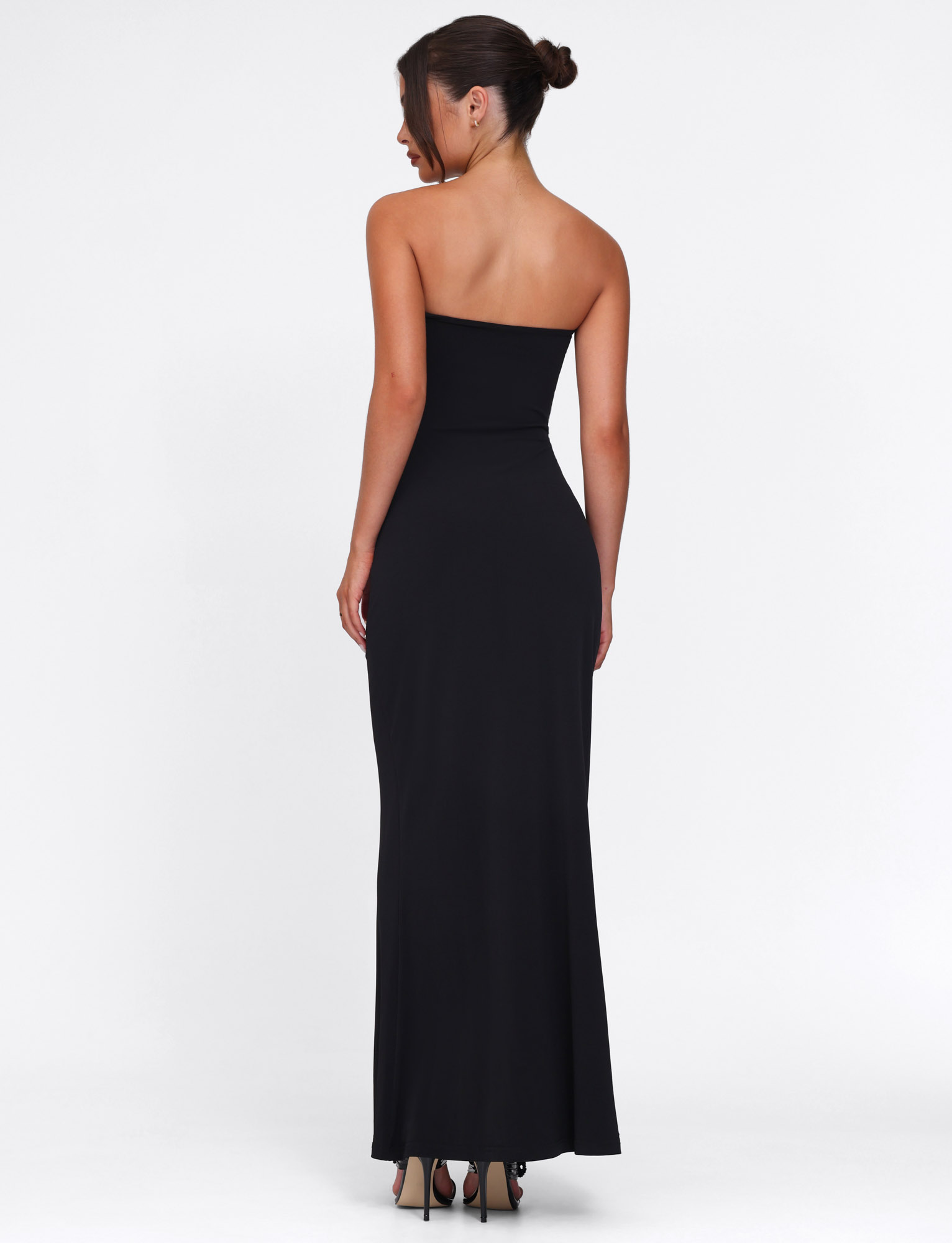 A woman with her hair in a bun models the TIGERMIST CECILY MAXI DRESS - BLACK, a strapless maxi with a sweetheart neckline, paired with high heels as she stands facing away from the camera against a plain white background.