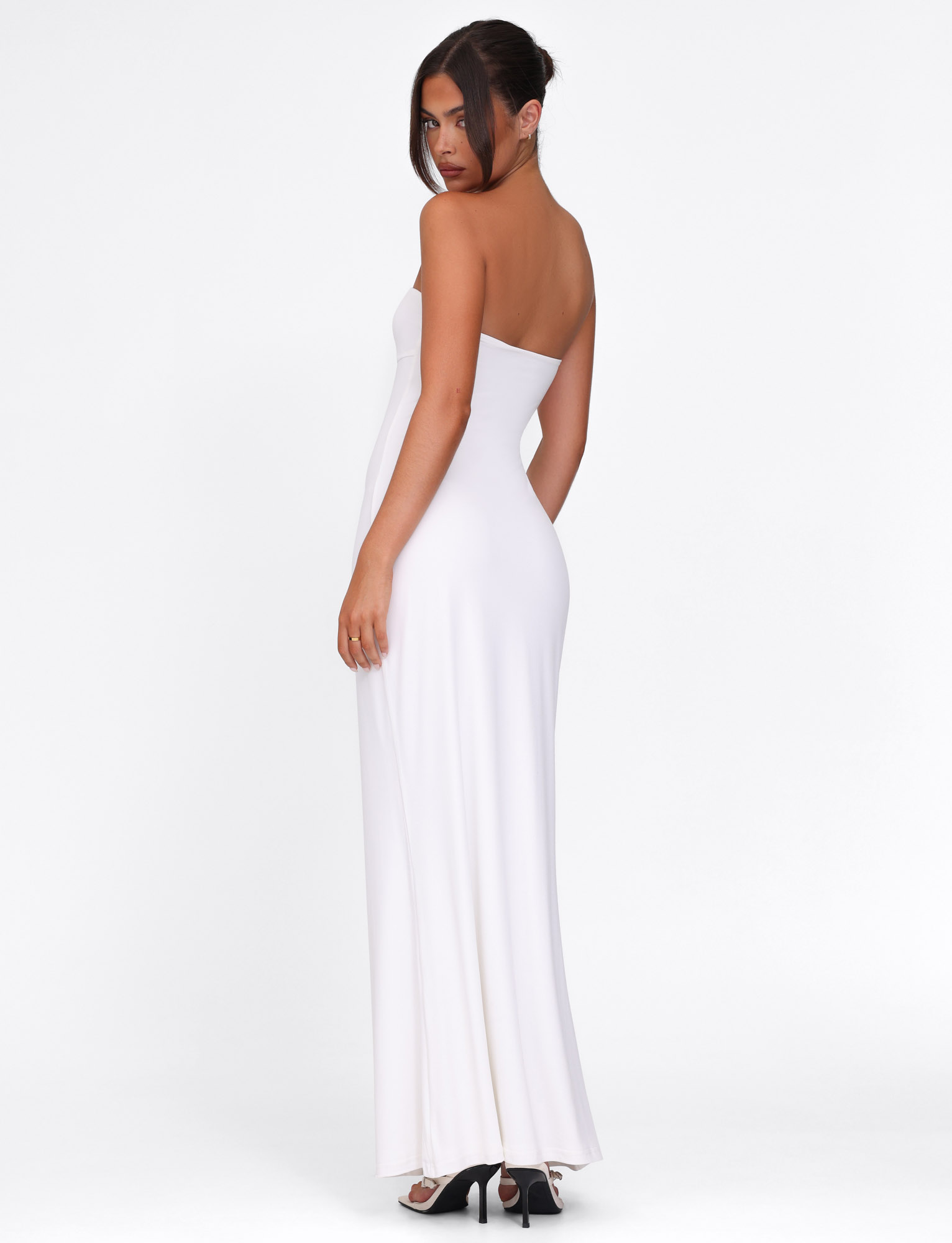 A woman with dark hair in an updo wears the TIGERMIST CECILY MAXI DRESS in white—a fitted, strapless silhouette—paired with black heels, standing and looking over her shoulder against a plain white background.