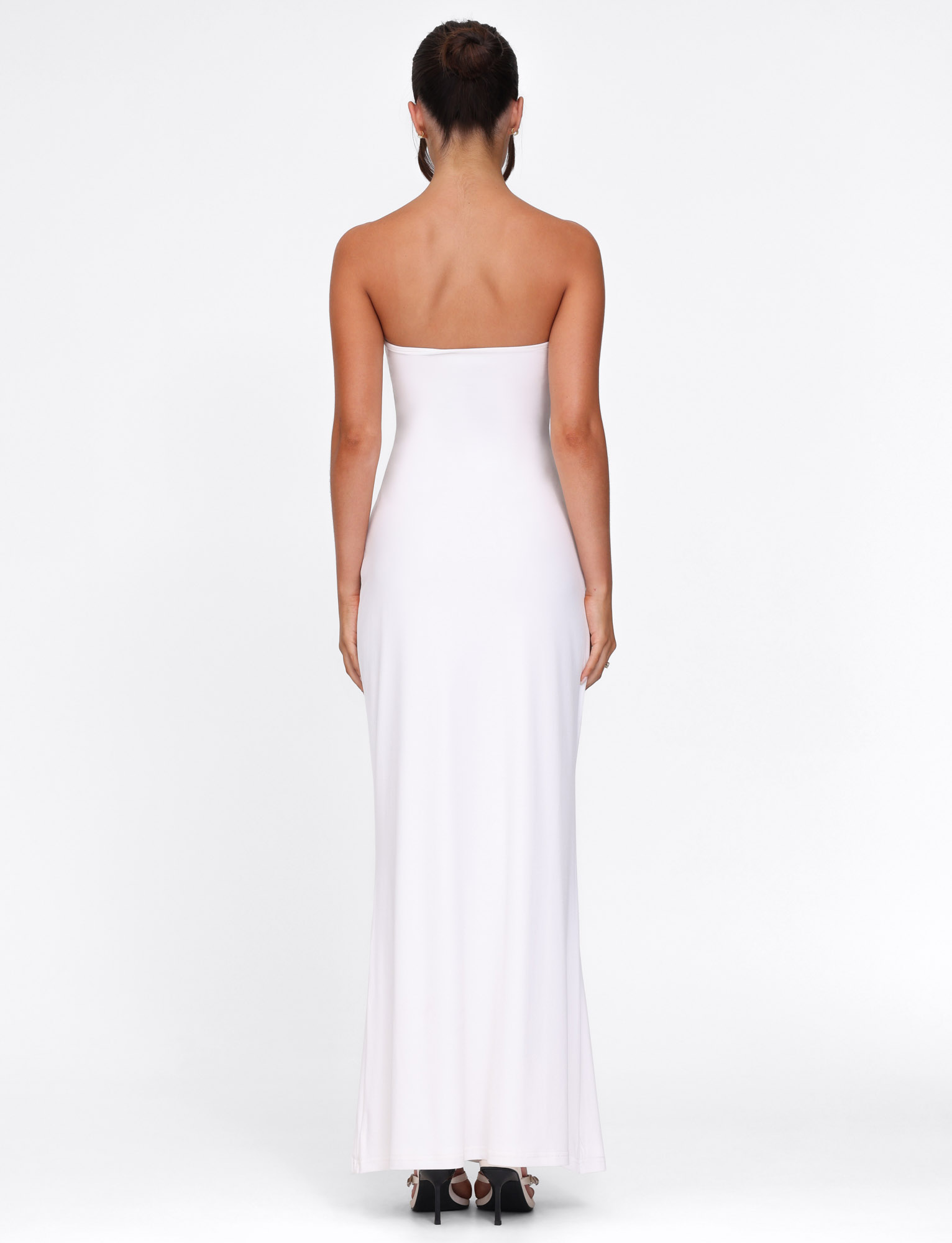 A woman stands facing away, wearing the TIGERMIST CECILY MAXI DRESS in white. This strapless, fitted silhouette maxi dress is paired with black open-toe heels. Her hair is styled up, and the background is plain white.