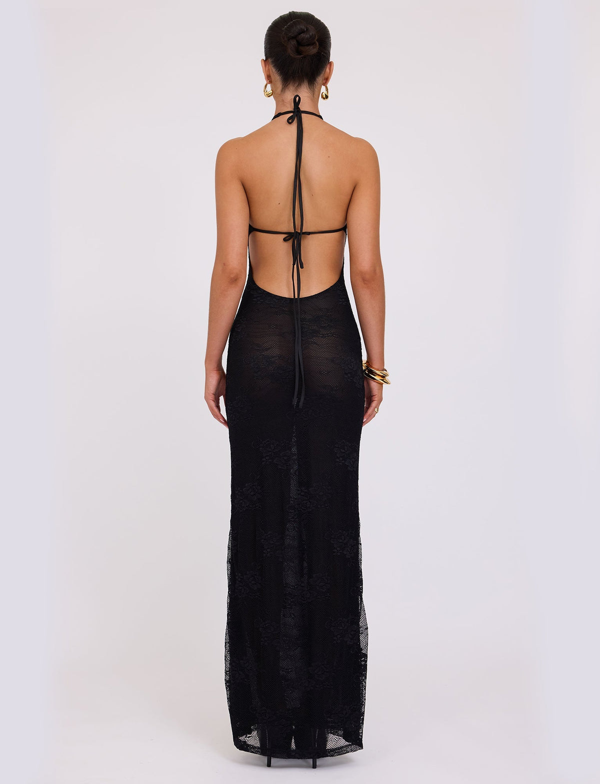 A woman stands with her back to the camera, wearing the LUCIA DRESS - BLACK : LACE from TIGERMIST. The black halter-neck gown features an open back secured with thin straps, and she has styled her hair in a bun. Her ensemble is complemented by gold hoop earrings and gold bangles. The floor-length dress is slightly transparent.