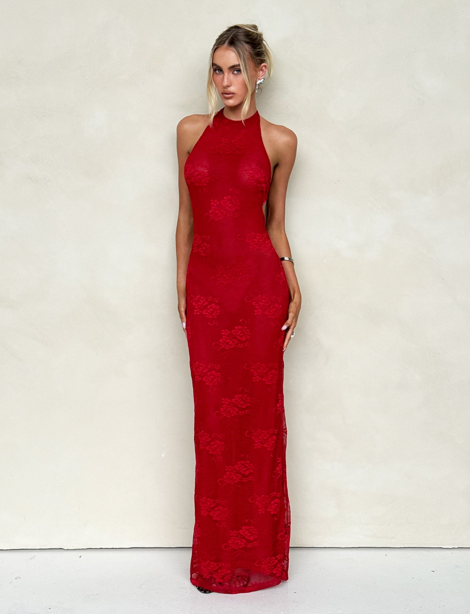 A woman stands against a plain wall, wearing the form-fitting LUCIA DRESS - RED : LACE from TIGERMIST that extends to the floor. She has her hair styled up and gazes directly at the camera.
