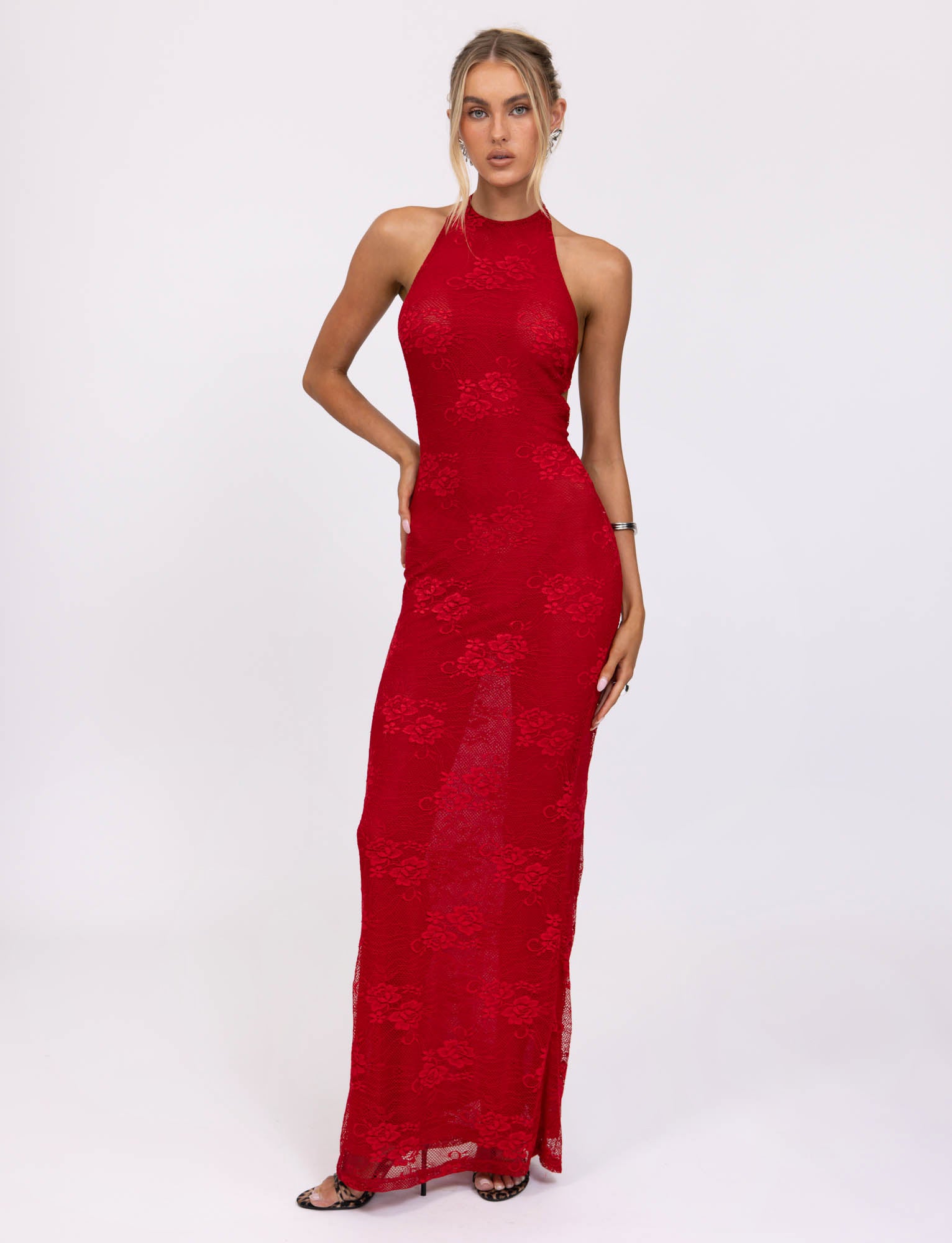 A woman is dressed in the LUCIA DRESS - RED : LACE from TIGERMIST, a sleeveless, form-fitting gown that reaches the floor. She poses against a plain white background with one hand on her hip and the other resting by her side. Her outfit is accessorized with bracelets and she wears black high heels.