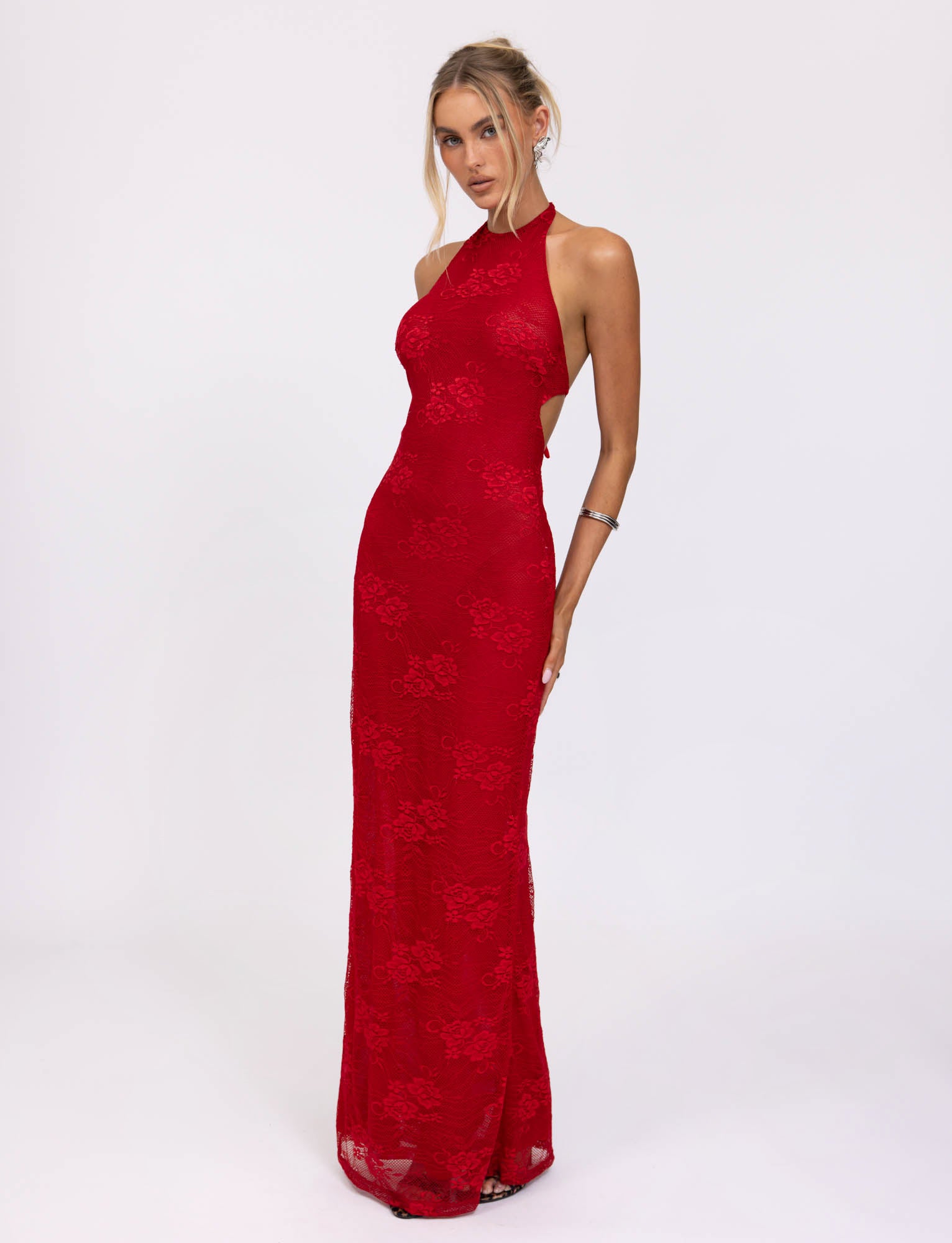 A woman in the elegant Lucia Dress from Tigermist stands against a plain white background. The red lace dress is form-fitting and floor-length, accentuating her figure. She has her hair styled in a loose updo and wears minimal jewelry.