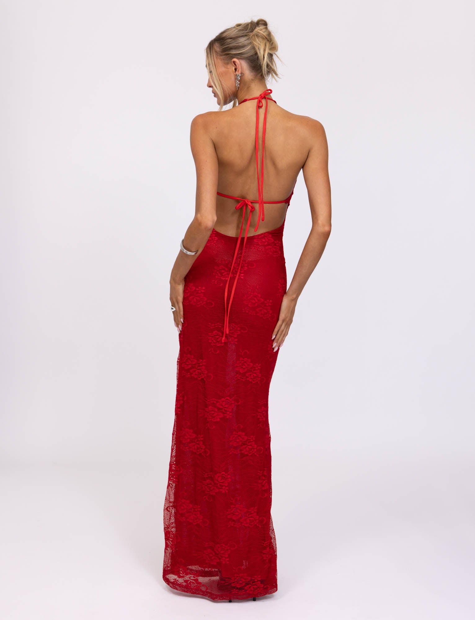 A person with blonde hair styled in an updo is wearing a stunning TIGERMIST LUCIA DRESS - RED : LACE. The dress features a long, fitted silhouette with tied straps at the neck and waist, beautifully showcasing their back against a plain white background.