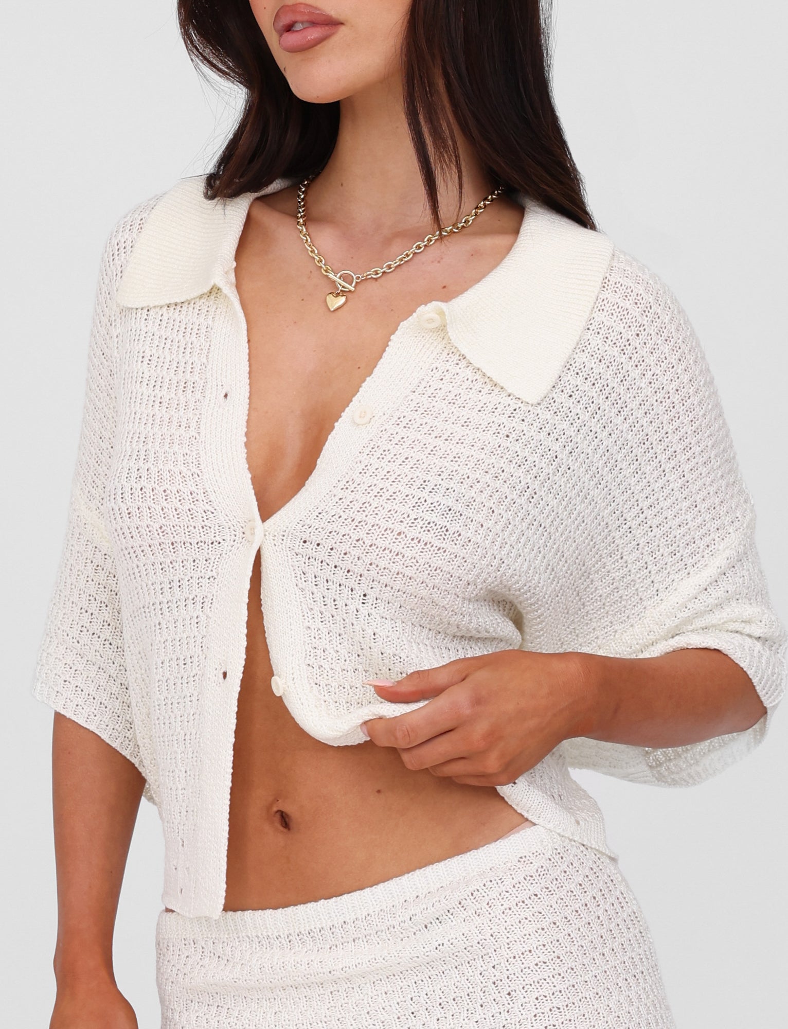 A woman with long dark hair wearing the oversized fit JANAI TOP - WHITE : CREAM from TIGERMIST, along with a heart-shaped necklace. The partially buttoned, collared neckline cardigan is held gently as she tugs at its hem, and it’s paired perfectly with a matching knit skirt.