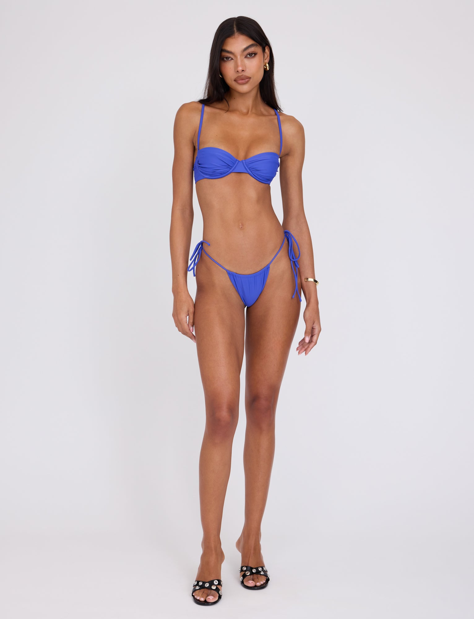 A woman stands against a plain white background, wearing the TIGERMIST KHESSA BIKINI TOP in cobalt blue. She has long dark hair and is looking directly at the camera. She is also wearing black sandals and a gold bracelet on her left wrist.