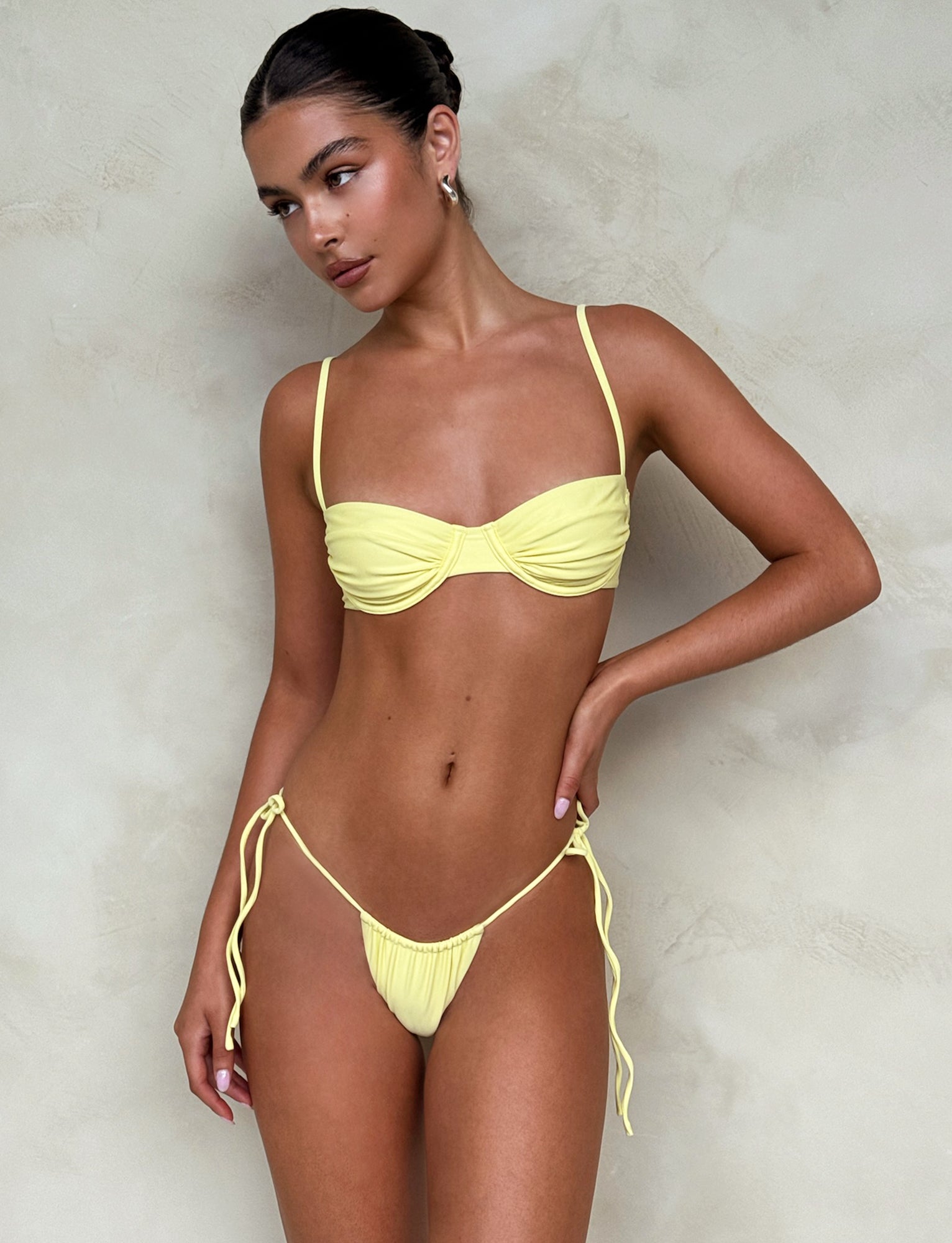 A woman stands against a textured beige background, wearing the KHESSA BIKINI BOTTOM - YELLOW: LEMON from TIGERMIST. She has her hair pulled back and is looking to her right. She accessorizes with small hoop earrings and exudes confidence and relaxation.