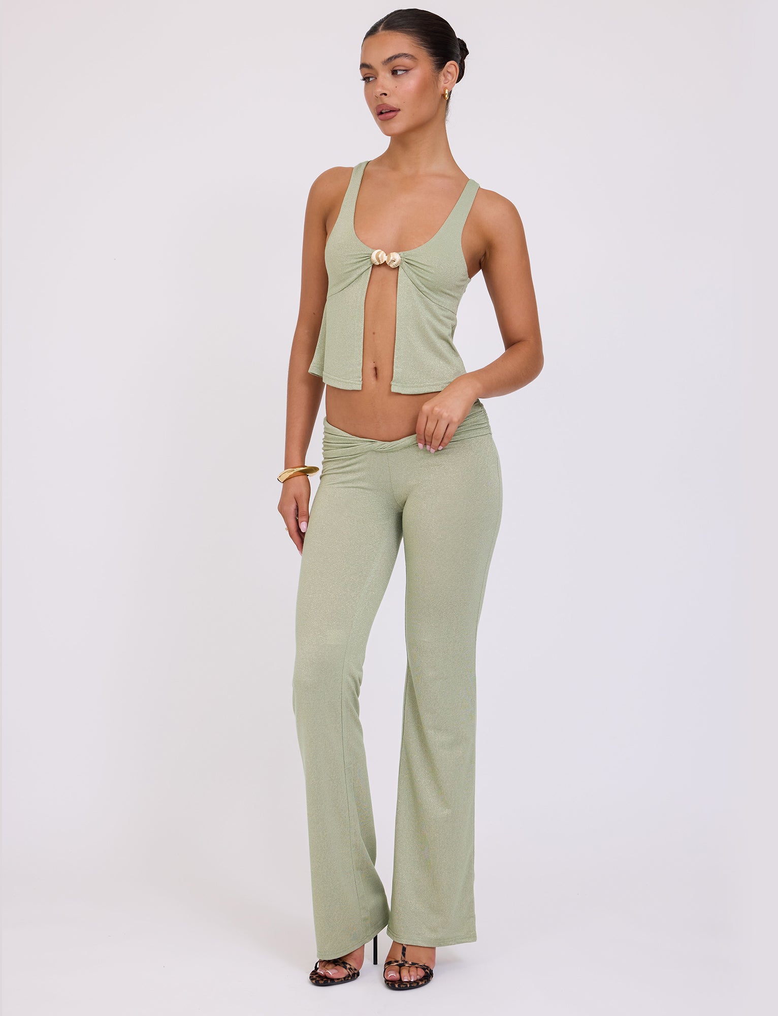 A woman stands posing against a plain white background, showcasing a chic Tiger Mist ensemble. She is wearing the Amethyst Pant in Green Sparkle, paired with a coordinating light green, sleeveless top featuring a front tie. She accessorizes with gold bracelets and black strappy heels. Her hair is neatly styled in an updo.