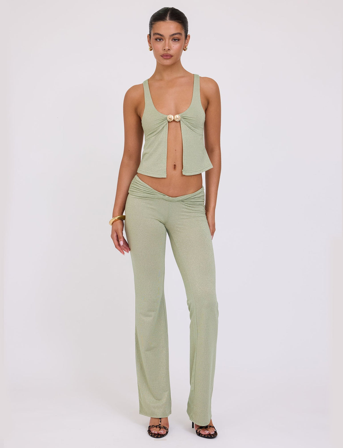 A woman stands against a plain white background, wearing the AMETHYST PANT - GREEN SPARKLE from TIGERMIST. The top features a deep neckline and front tie detail, while the high-waisted pants are flared. She accessorizes with a gold bracelet and black heeled sandals.