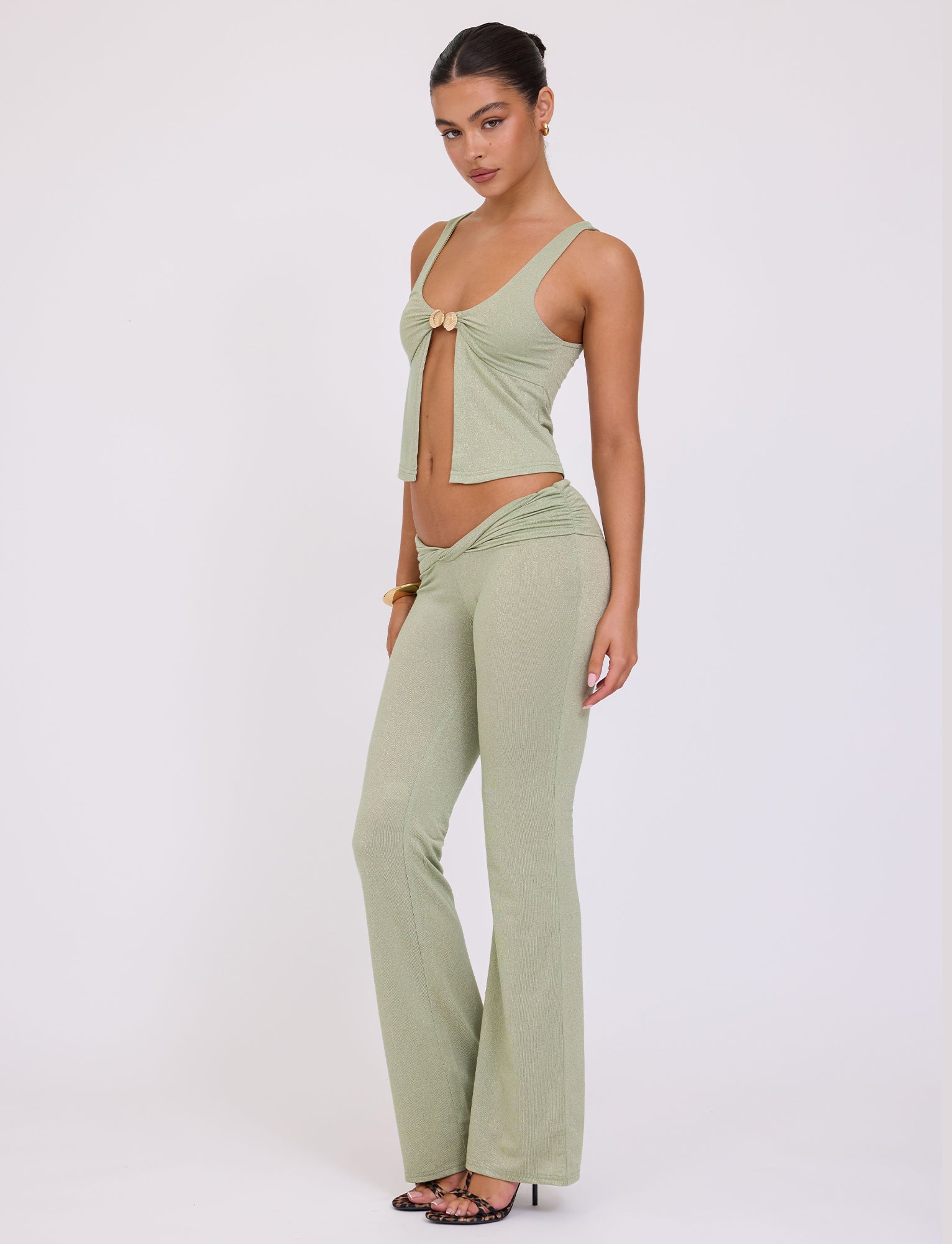 A woman stands against a plain white background, wearing the chic green sparkle Amethyst Top by TigerMist. Her stylish ensemble includes a sleeveless top with a front tie and high-waisted, flared pants. With her hair pulled back and black strappy heels, she effortlessly exudes chic elegance.