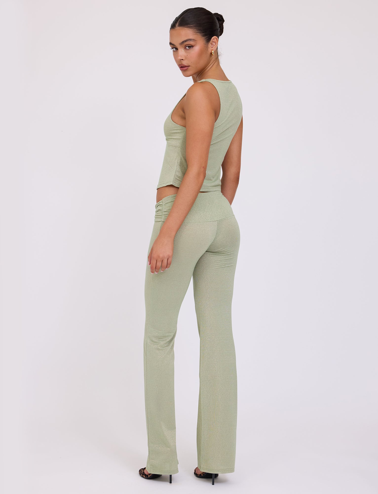A woman with dark hair in a sleek bun is wearing the AMETHYST TOP - GREEN : GREEN SPARKLE from TIGERMIST along with matching high-waisted pants. She is standing sideways, turning her head slightly to look over her shoulder, against a plain white background.