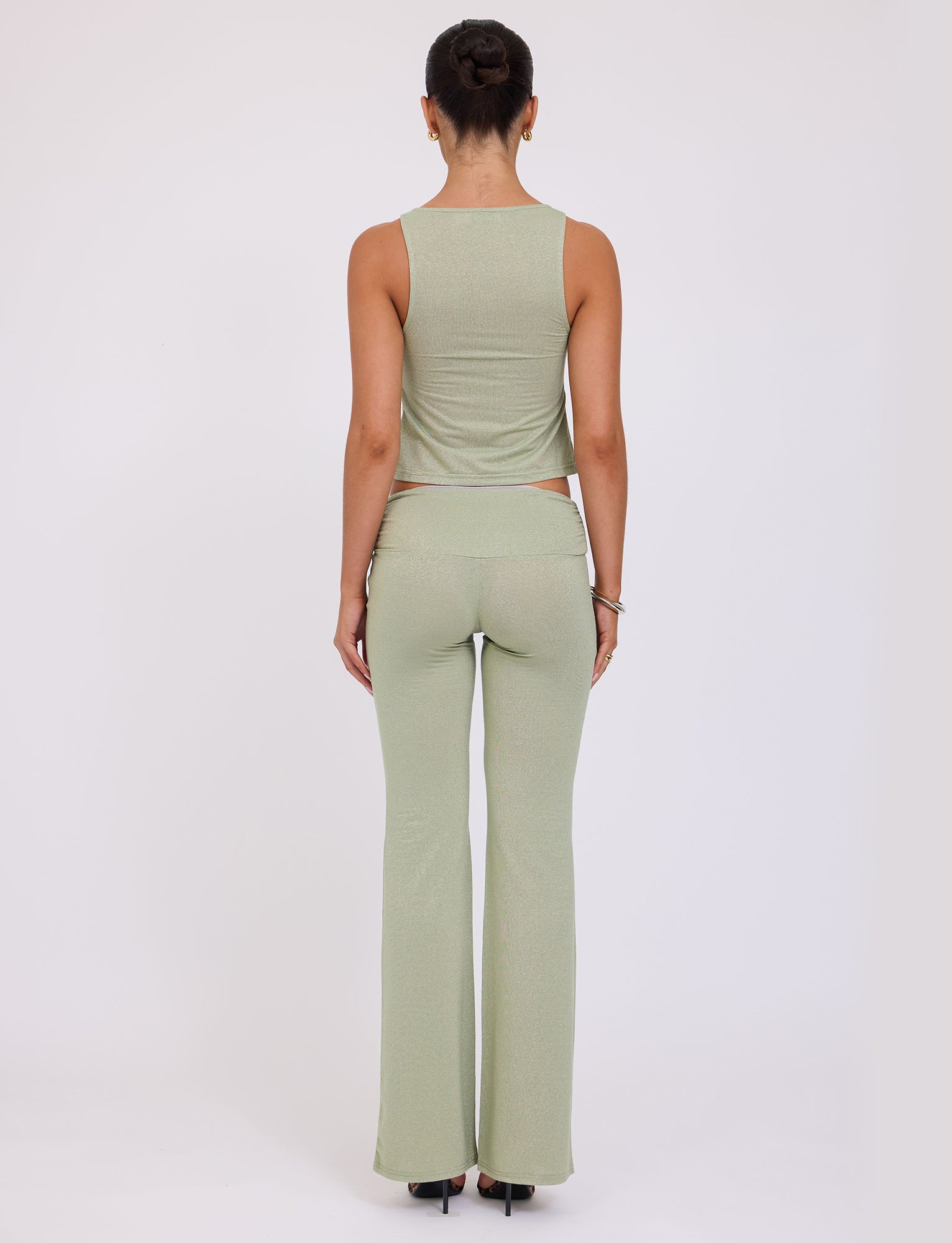 A person is seen from the back, standing with their hair in a bun. They are wearing a light green sleeveless top and matching AMETHYST PANT - GREEN : GREEN SPARKLE from TIGERMIST, along with black heels. The background is a plain, light-colored surface.