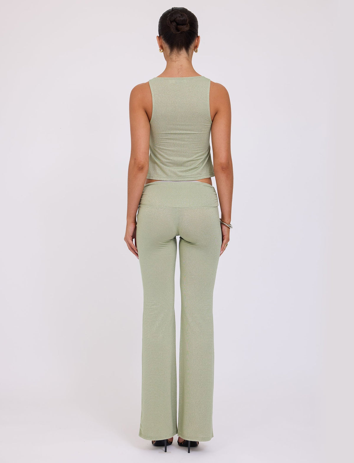 A person is seen from the back, standing with their hair in a bun. They are wearing a light green sleeveless top and matching AMETHYST PANT - GREEN : GREEN SPARKLE from TIGERMIST, along with black heels. The background is a plain, light-colored surface.