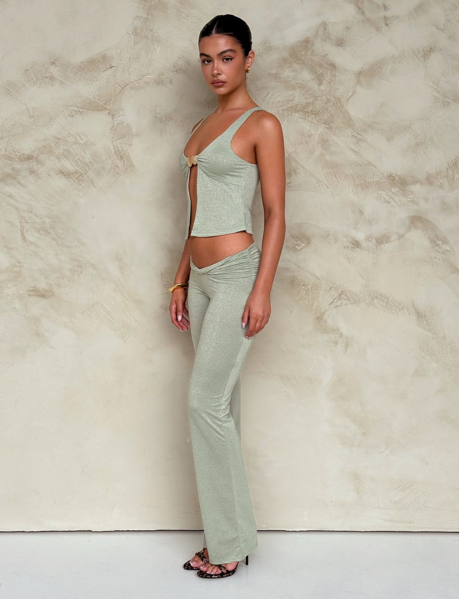 A woman poses against a textured beige wall, wearing the AMETHYST PANT - GREEN : GREEN SPARKLE from TIGERMIST, featuring a light green sleeveless crop top with a front tie and matching high-waisted pants. She has long dark hair tied back, minimal makeup, and wears black strappy heels along with a bracelet and earrings.