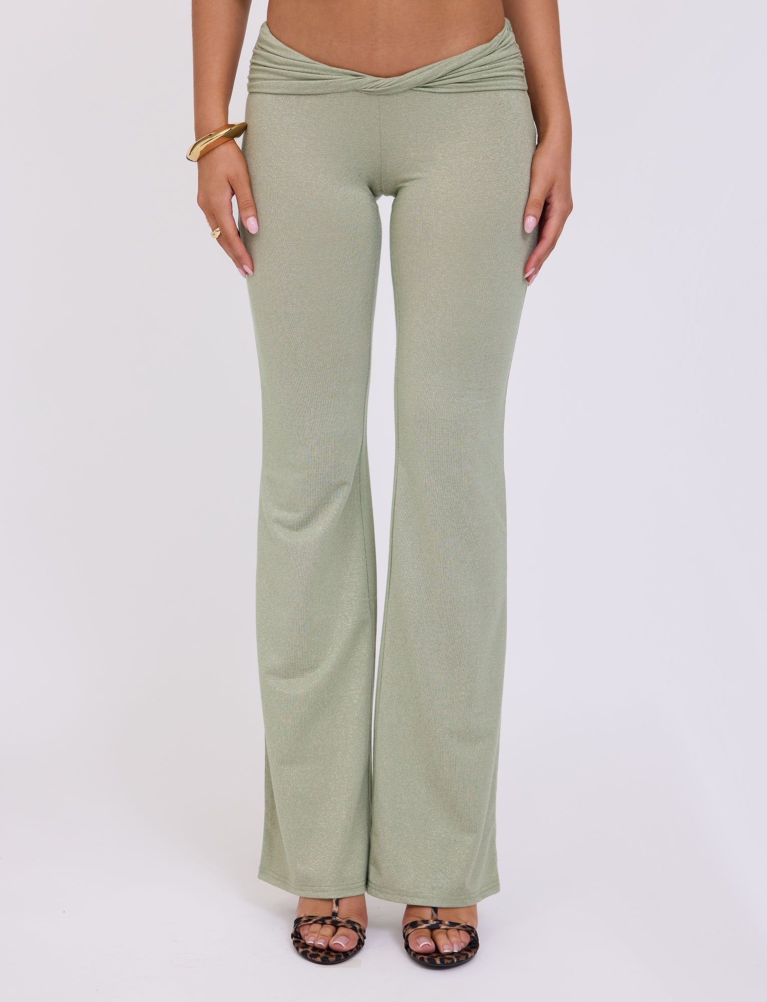 A person wearing the AMETHYST PANT - GREEN in GREEN SPARKLE by TIGERMIST, paired with black strappy heels. They have a gold bracelet on their right wrist. The background is plain white.