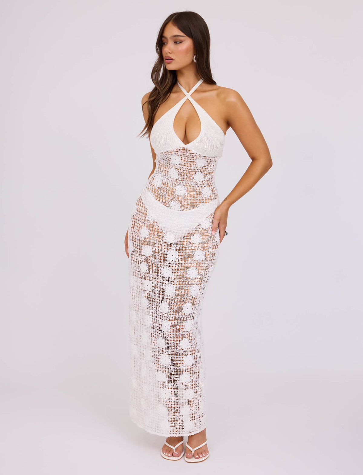 A woman is wearing the ARDEEN DRESS - WHITE from TIGERMIST, which boasts a detailed open-knit design. This sleeveless crochet dress features a halter neckline and a long, form-fitting silhouette. She has paired it with white strappy sandals and is posing against a plain light-gray background.