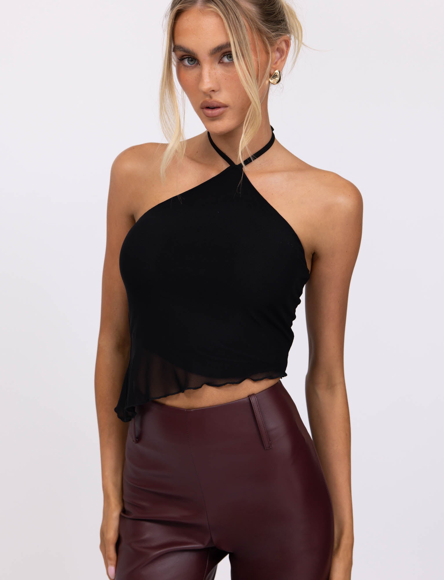 A woman with blonde hair styled in a loose updo wears the HESTIA TOP - BLACK, a sleek mesh halter top, and high-waisted burgundy leather pants, showcasing a fitted silhouette. She accessorizes with hoop earrings and several rings on her fingers. Standing against a neutral, pale background, she rests her hands on her thighs.