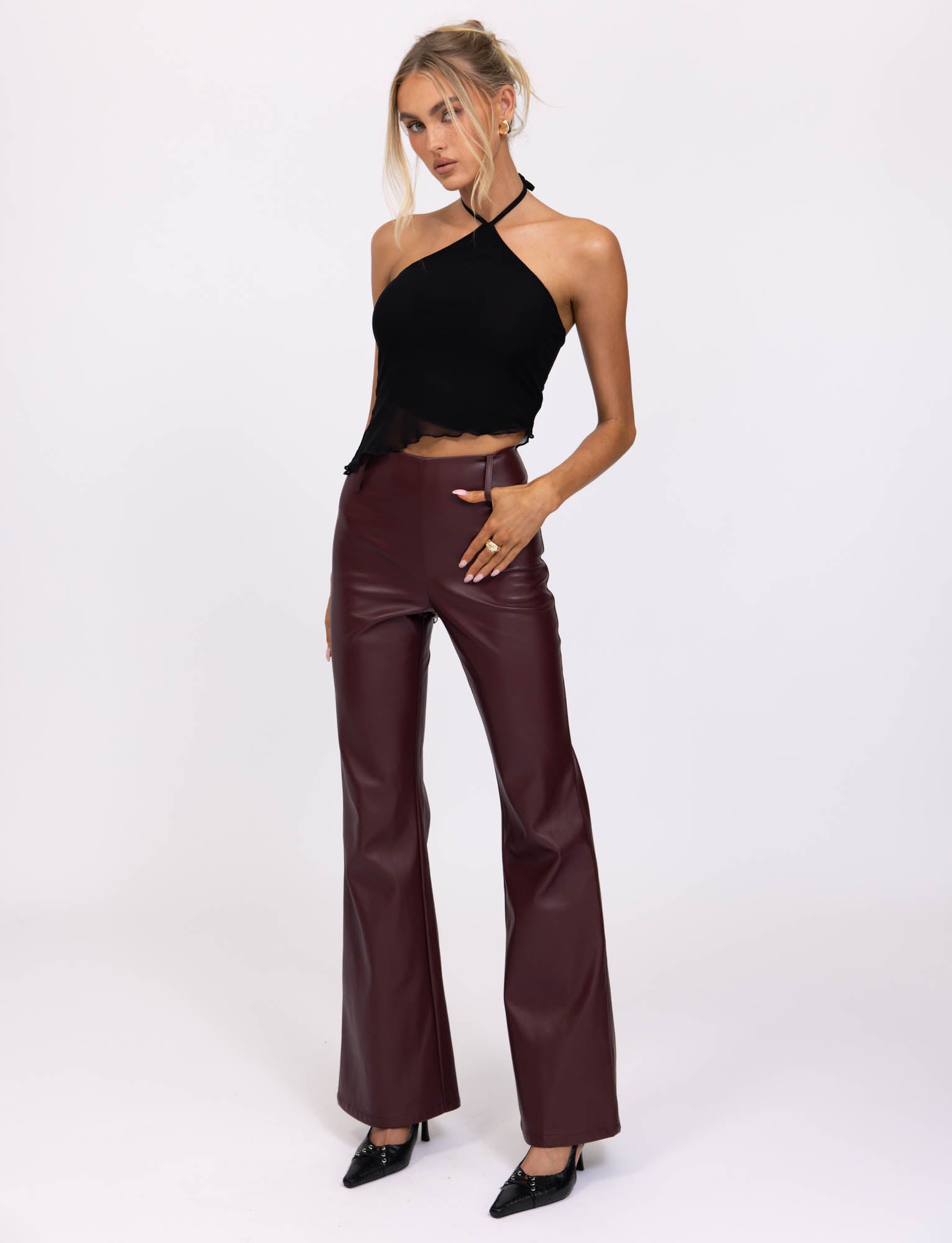 A woman with blonde hair styled in a loose updo wears the HESTIA TOP - BLACK, a sleek mesh halter top, and high-waisted burgundy leather pants, showcasing a fitted silhouette. She accessorizes with hoop earrings and several rings on her fingers. Standing against a neutral, pale background, she rests her hands on her thighs.
