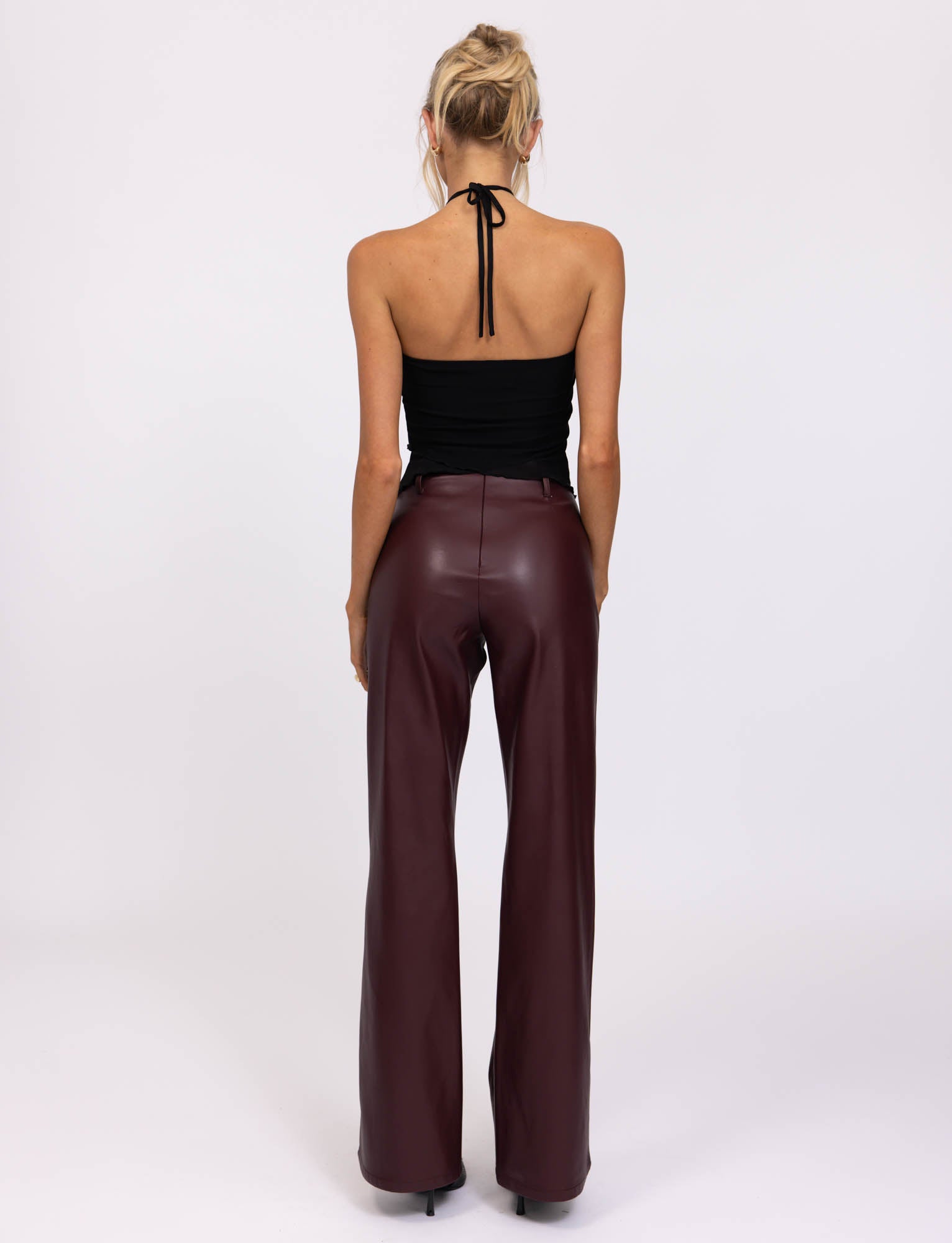 A woman with blonde hair styled in a loose updo wears the HESTIA TOP - BLACK, a sleek mesh halter top, and high-waisted burgundy leather pants, showcasing a fitted silhouette. She accessorizes with hoop earrings and several rings on her fingers. Standing against a neutral, pale background, she rests her hands on her thighs.