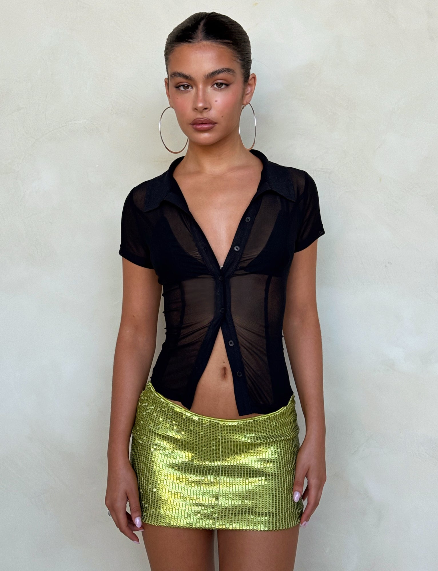 A person stands in front of a neutral background wearing the TIGERMIST AHLING TOP in black with short sleeves and a green sequin mini skirt. They have large hoop earrings and a neutral expression. Their hair is pulled back tightly.