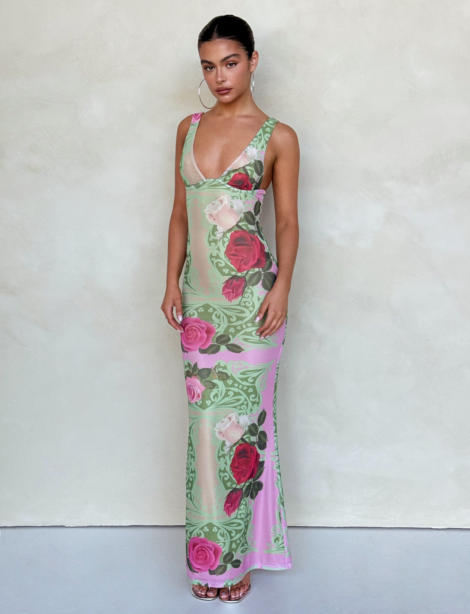 A woman stands against a plain backdrop, wearing the LUNARIA DRESS - MULTI : MACRO FLORAL : SUMMER ROSE from TIGERMIST. The long, sleeveless dress features a deep V-neckline and a light green and pink floral pattern with various roses. She accessorizes with large hoop earrings and gold open-toe heels.