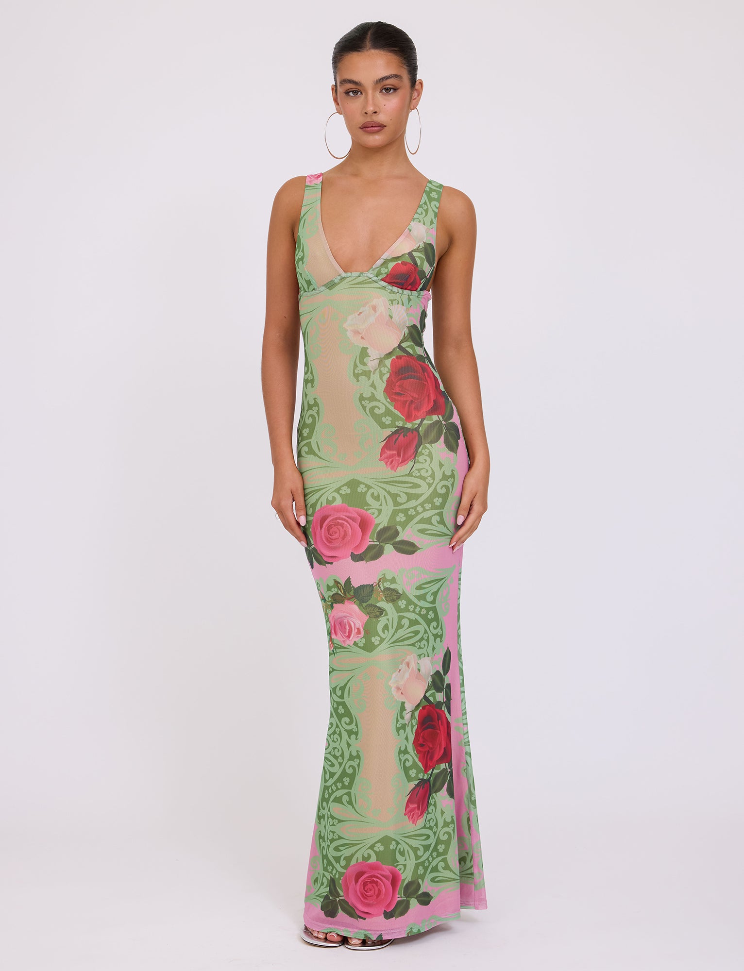 A woman stands against a plain white background wearing the LUNARIA DRESS - MULTI : MACRO FLORAL : SUMMER ROSE by TIGERMIST, a long, sleeveless dress with a plunging neckline. The dress showcases a pink floral pattern featuring roses and green leafy designs. She accessorizes with large hoop earrings and has her dark hair pulled back.