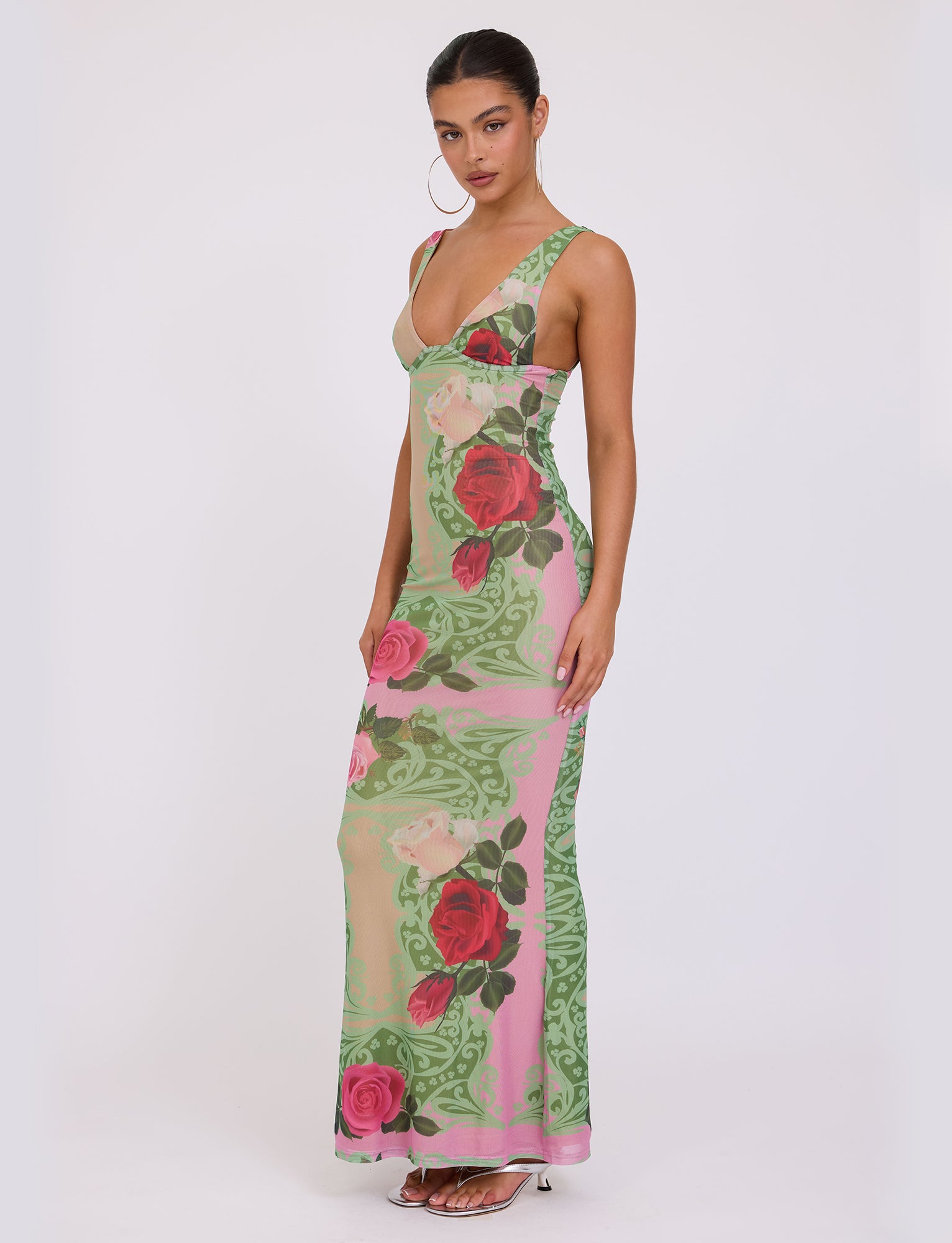 A woman stands against a plain background wearing the elegant, form-fitting LUNARIA DRESS - MULTI: MACRO FLORAL: SUMMER ROSE from TIGERMIST. Her hair is pulled back, and she accessorizes with large hoop earrings and strappy high-heeled sandals.