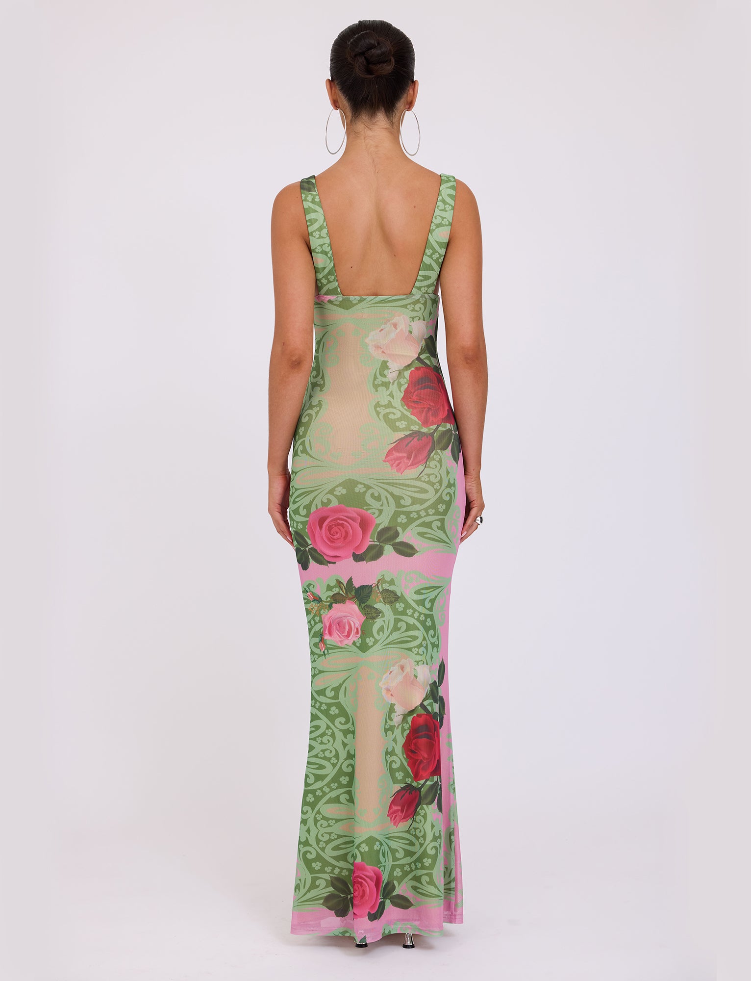 Facing away, a model showcases the LUNARIA DRESS - MULTI : MACRO FLORAL : SUMMER ROSE by TIGERMIST. This full-length, form-fitting dress is adorned with a vibrant floral pattern in shades of green, pink, and red. It features a low scoop back and wide shoulder straps. The model's hair is styled neatly in a bun.