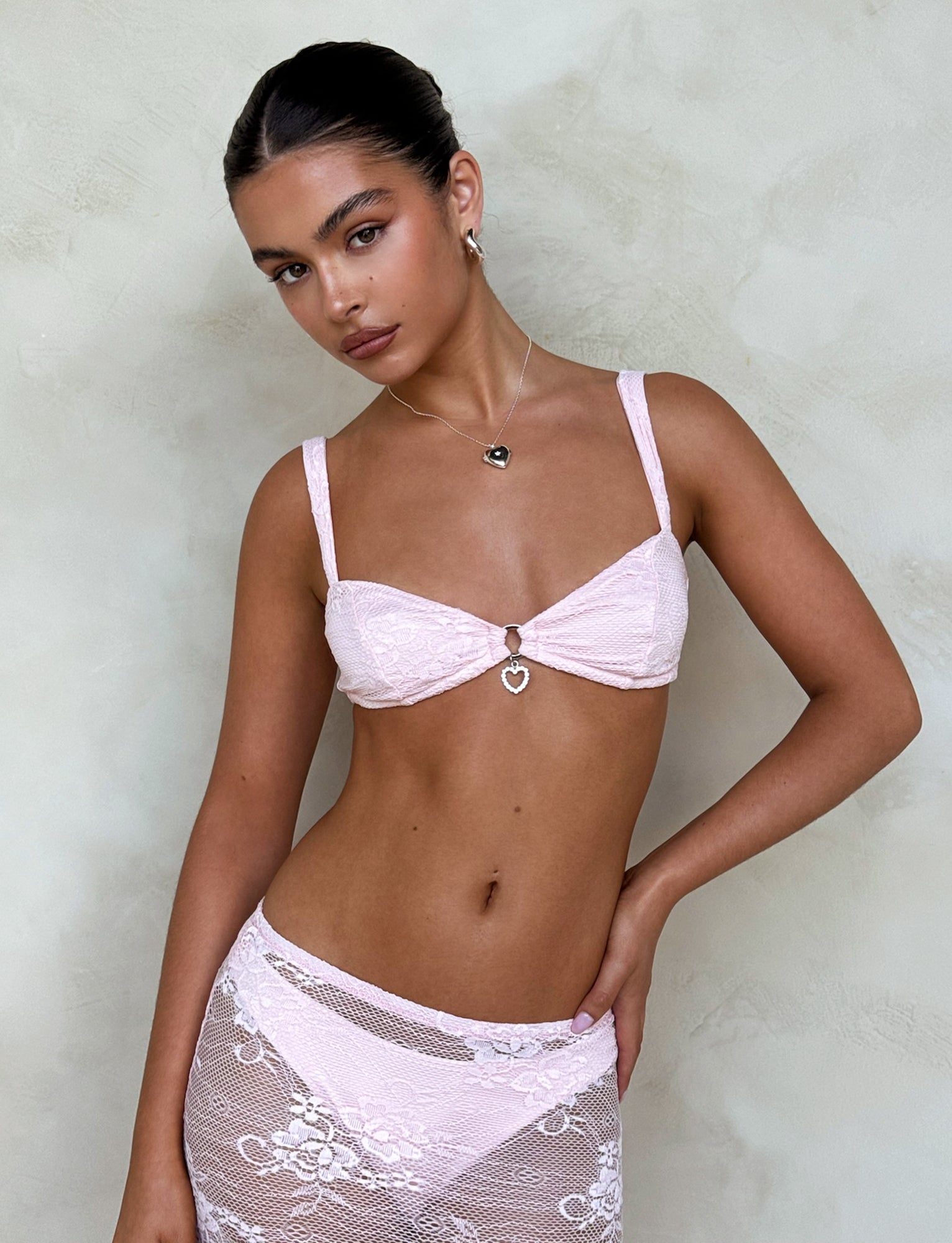 A woman is posing against a textured, neutral-colored wall, wearing a light pink Xadie Top by TIGERMIST featuring a heart-shaped charm in the center and a matching lace cover-up skirt. Her hair is tied back, and she accessorizes with a necklace and earrings.
