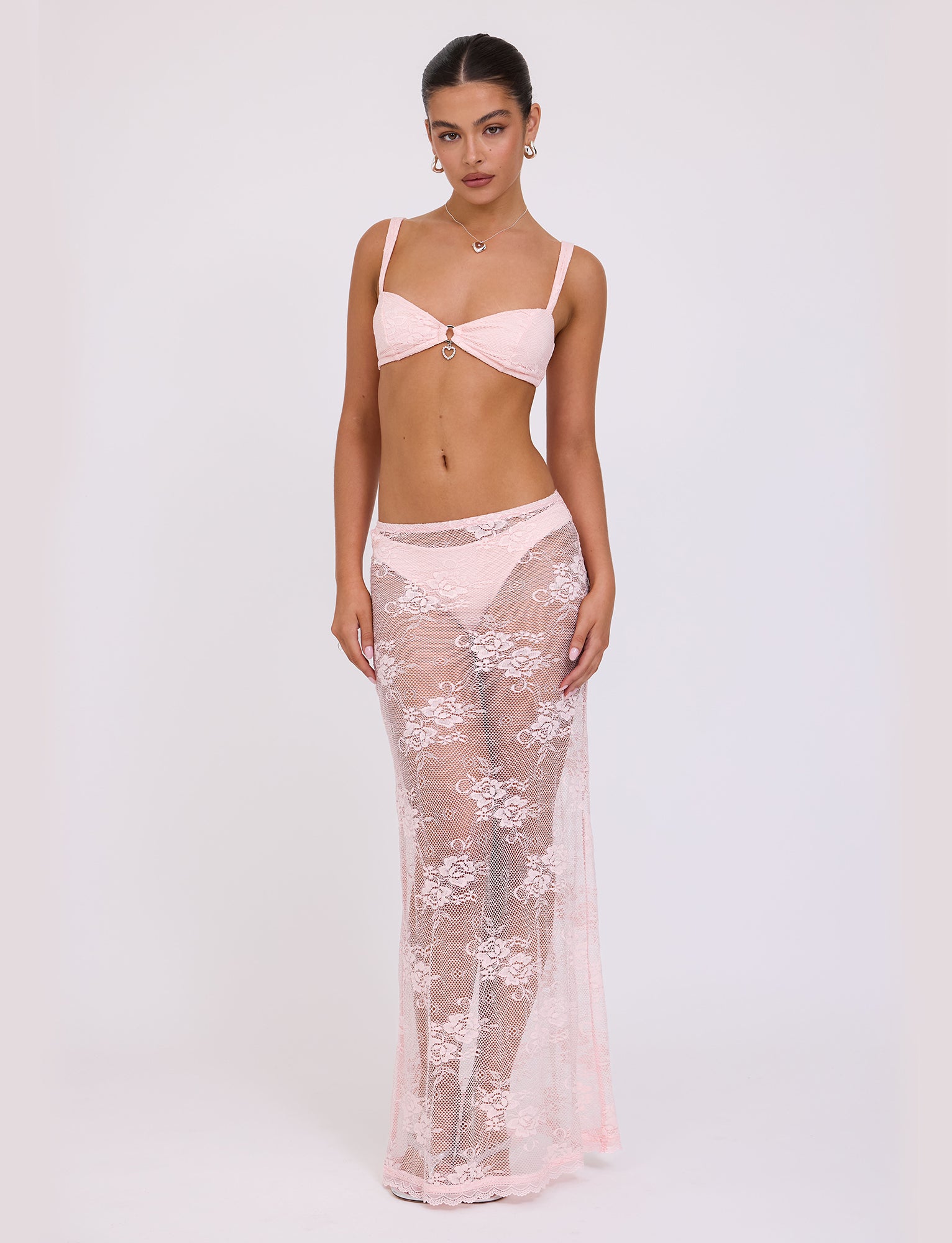 A woman stands against a plain background wearing the XADIE TOP in pale pink from TIGERMIST, paired with a matching sheer, floor-length lace skirt. She has long hair tied back and is accessorized with earrings and a necklace from Tiger Mist. She gazes directly into the camera with a neutral expression.