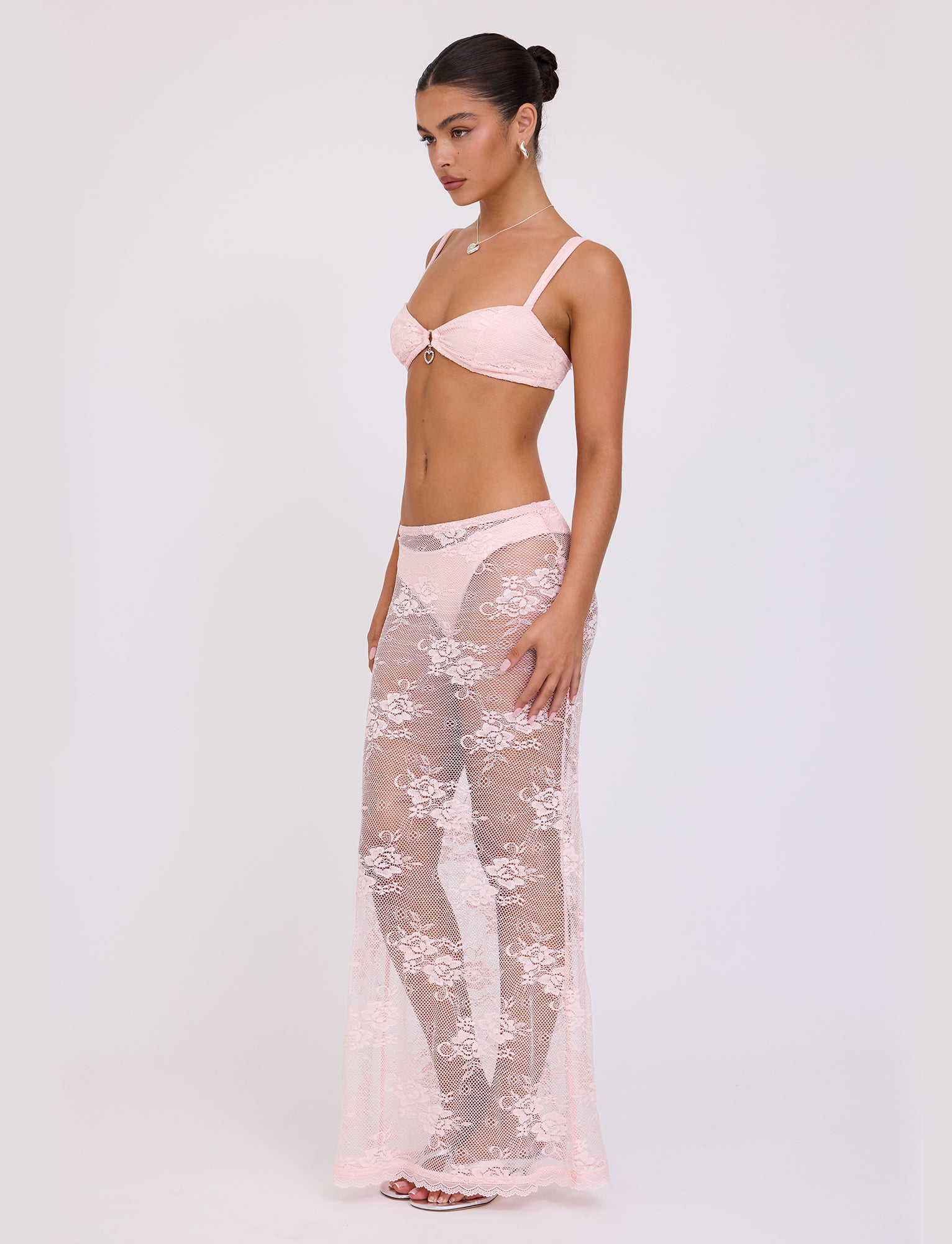 A woman stands against a plain background wearing the XADIE TOP from TIGERMIST in pastel pink. The outfit includes a bra, matching underwear, and a sheer lace skirt that extends to her feet. Her hair is styled neatly in a bun as she poses with her body turned slightly to the side.