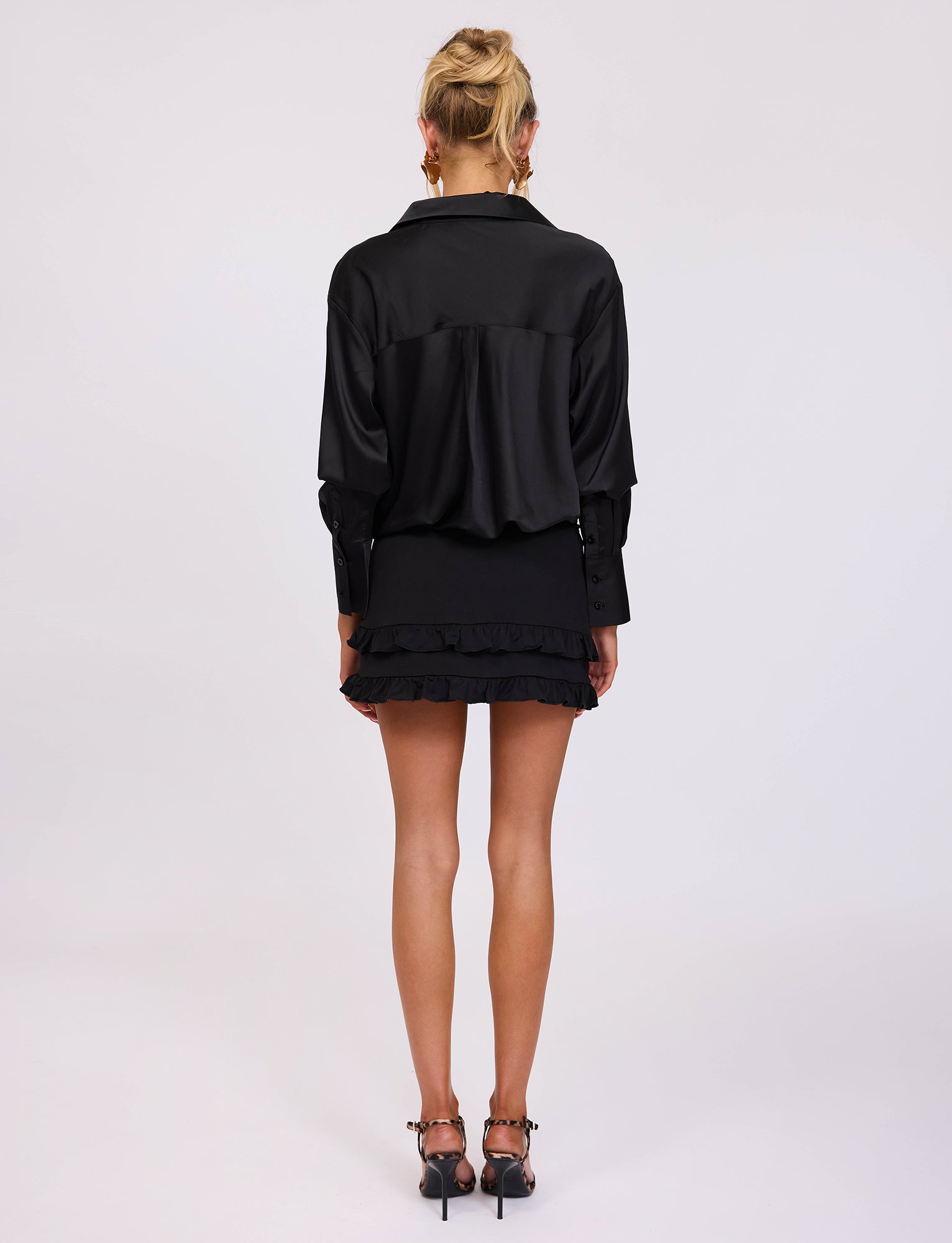 A person with blond hair stands facing away, wearing a chic TIGERMIST ALINA SHIRT in black and a short, ruffled black skirt. Their earrings glint under the light as they balance gracefully in black high-heeled shoes with decorative ankle straps. The background is plain white.