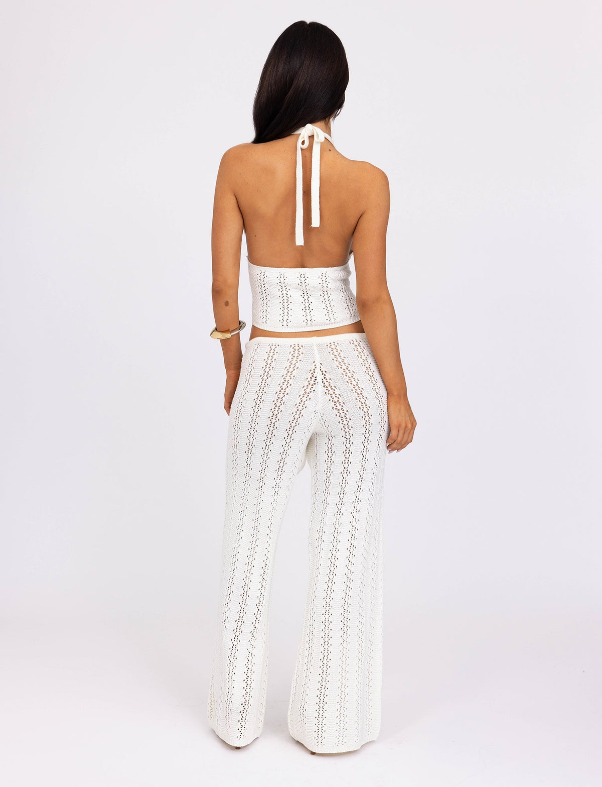A person with long dark hair, seen from the back, wears a white crochet halter top and the SANDOR PANT - WHITE from TIGERMIST. The top ties around the neck, and the sheer pants reveal a light-colored undergarment. The background is white.