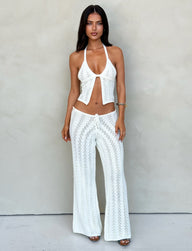 A woman stands in front of a neutral background wearing TIGERMIST's matching white crochet halter top and SANDOR PANT - WHITE. The halter top has an open front and ties at the neck, while the pants feature an intricate crochet pattern. She's accessorized with a bracelet.