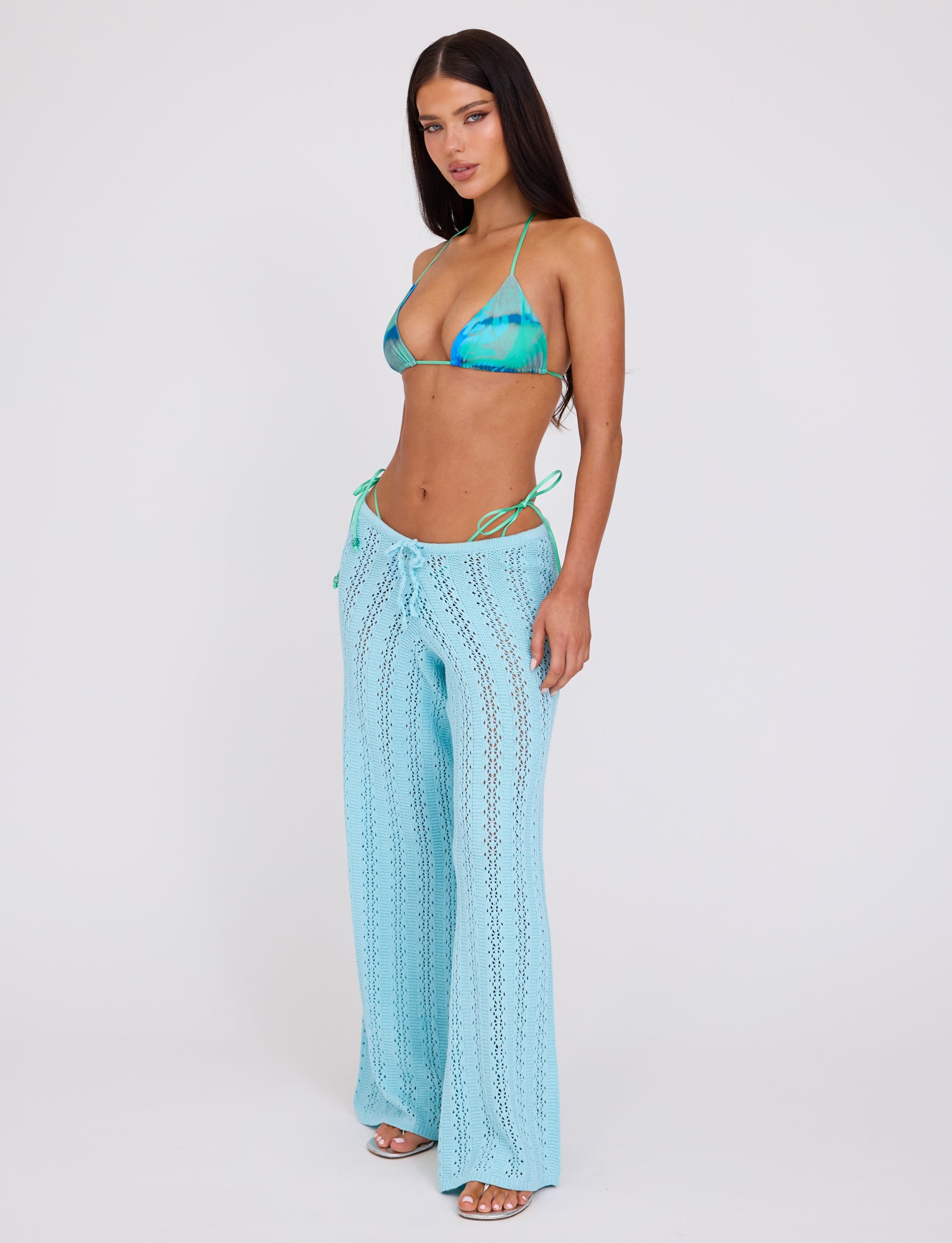 A woman with long dark hair wears a blue and green tie-dye bikini top from Tiger Mist and TIGERMIST's SANDOR PANT in BABY BLUE. She poses against a plain white background, standing with one hand on her hip, also wearing strappy high heels.